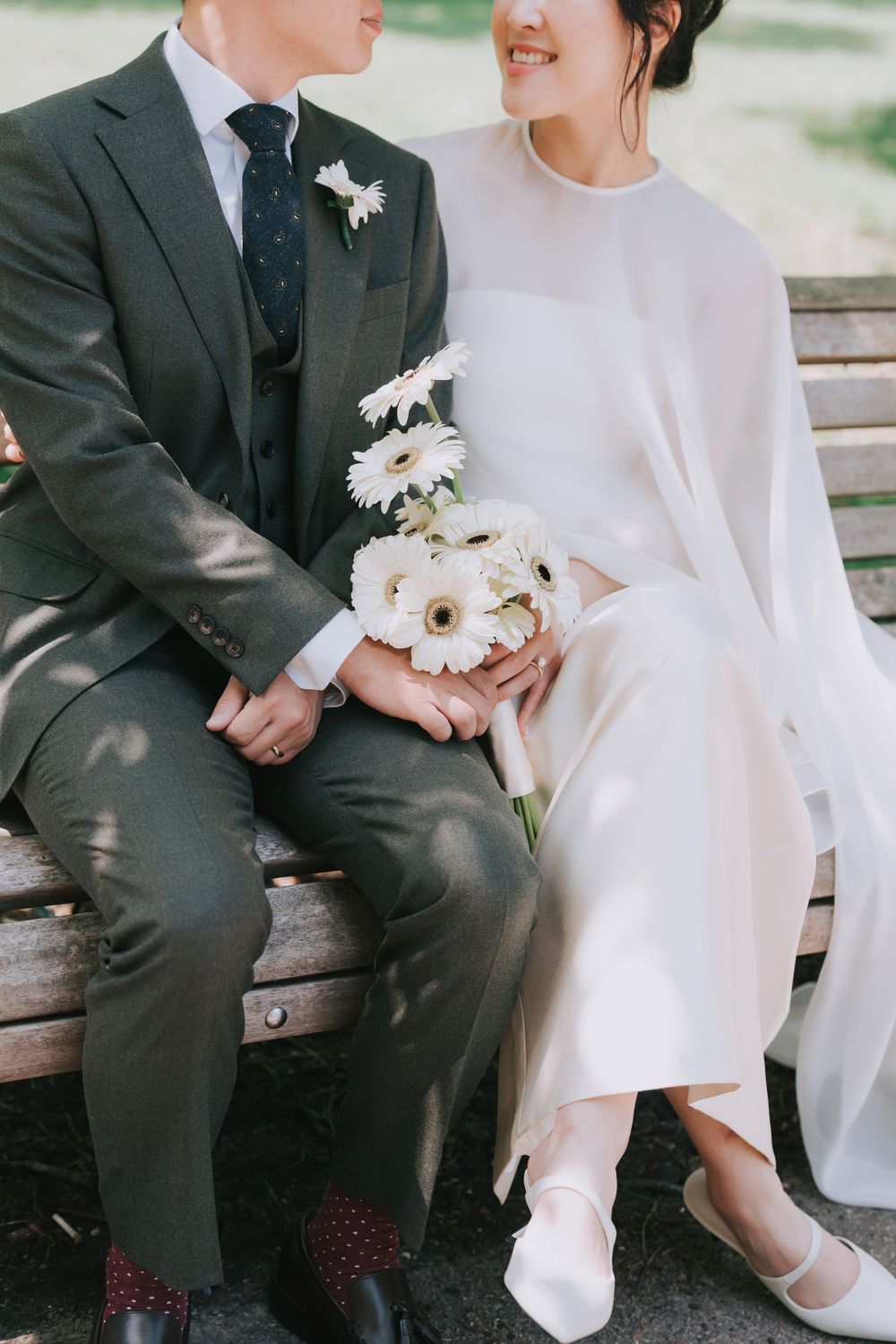 Bride and groom sitting on a bench, holding a white gerbera daisy bouquet, dressed in wedding attire.