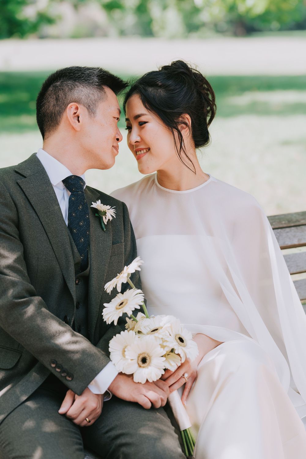 Asian bride and groom in elegant wedding attire sit on a bench, sharing an intimate moment with white gerbera bouquet.
