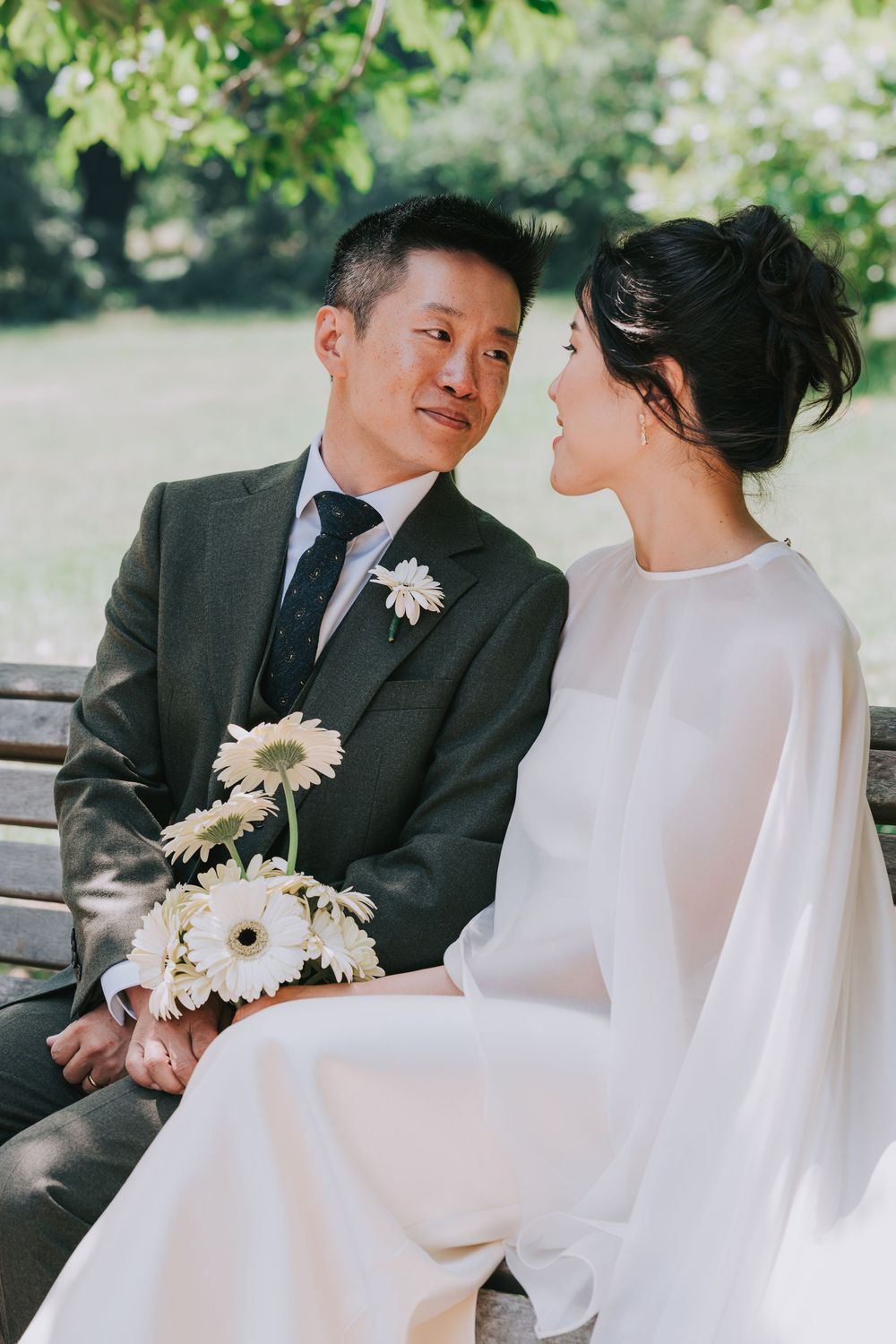 Asian couple in wedding attire sitting on a park bench, bride in white gown holding gerbera daisies, groom in gray suit.