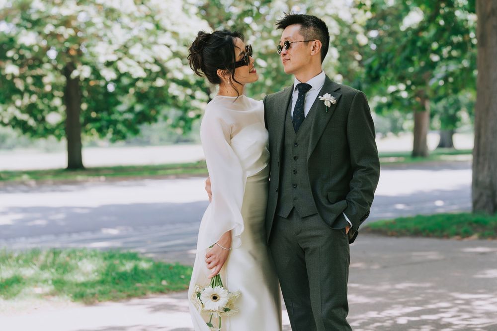 A bride and groom in elegant wedding attire pose together in a lush green park setting.