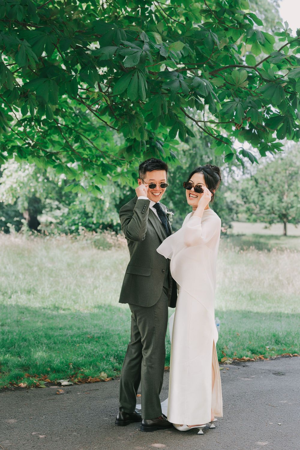Bride and groom posing under green tree in wedding attire wearing sunglasses outdoors.