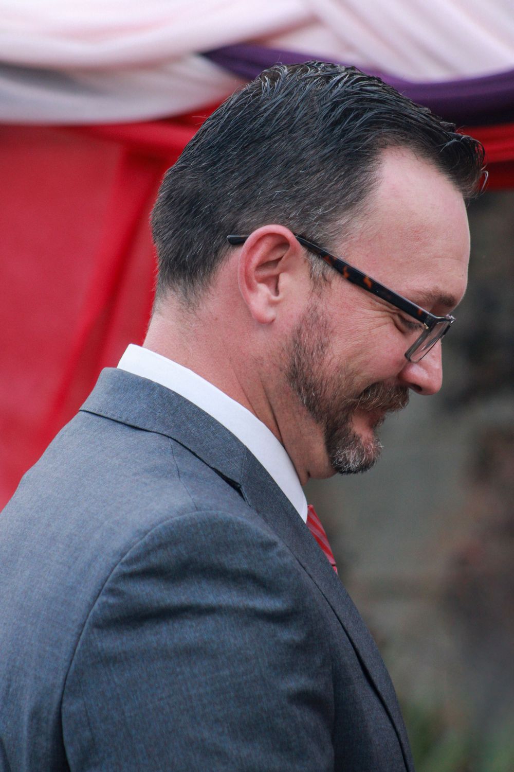 A man in a grey suit and glasses looks down while smiling at an outdoor event.