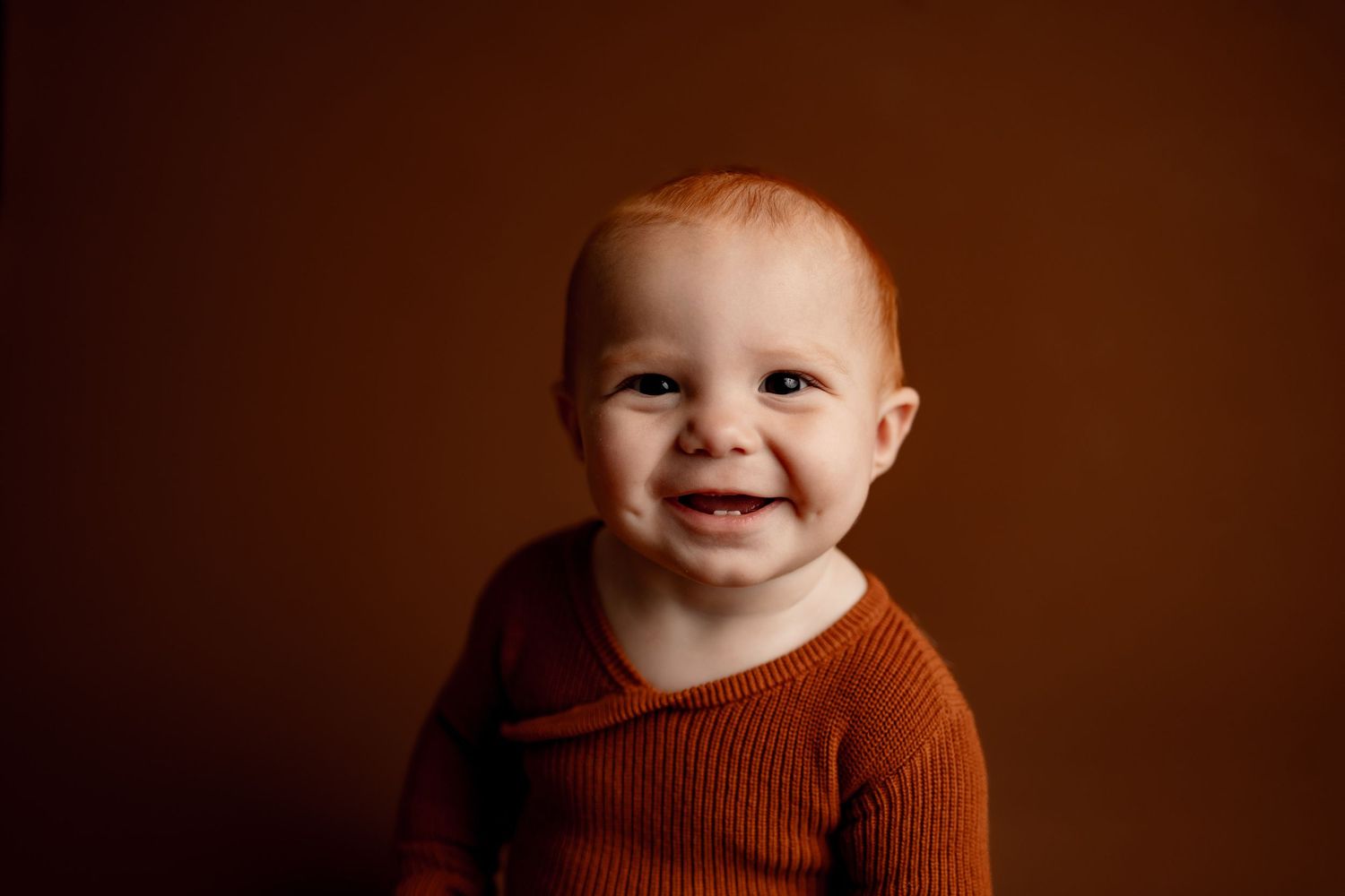 Red-haired baby in rust romper stands at small wooden stool with toy hourglass, brown studio backdrop.