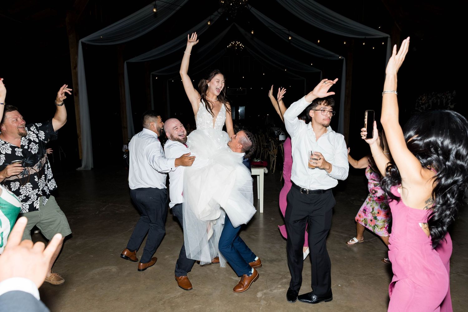 Guests celebrate at a lively wedding reception as the bride is lifted joyfully on the dance floor.