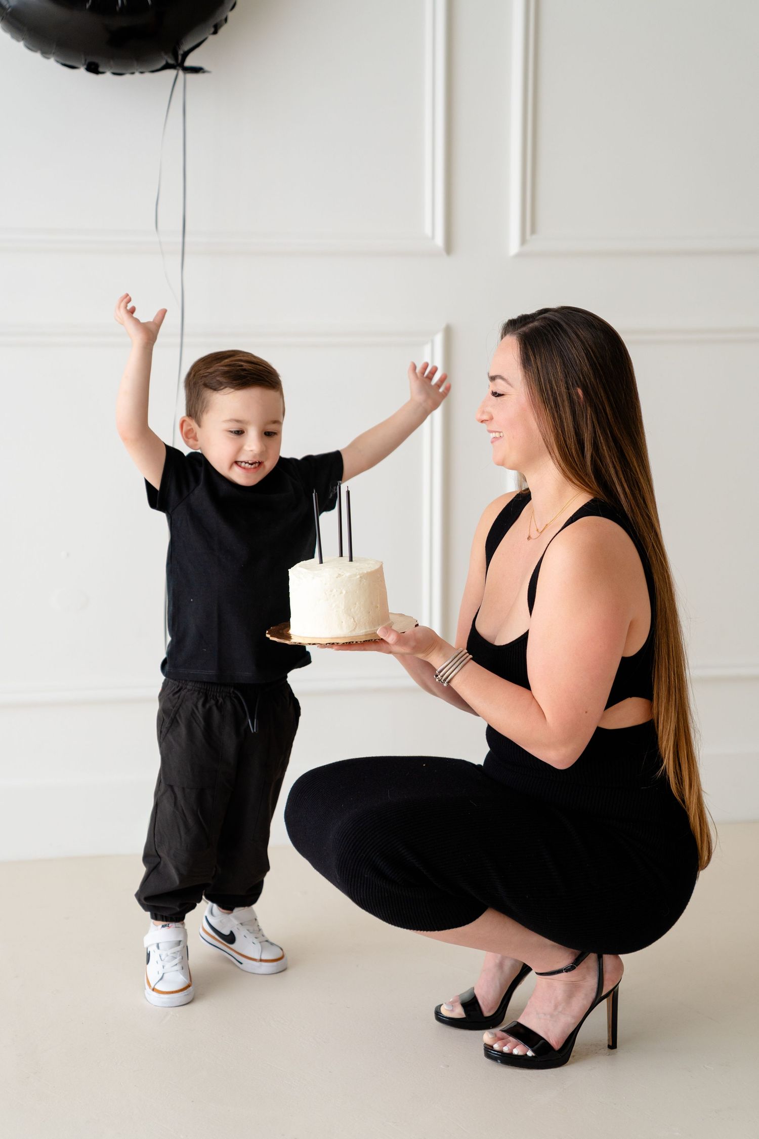 A woman in black holds a birthday cake while a young boy in black raises his arms in excitement near black balloons.