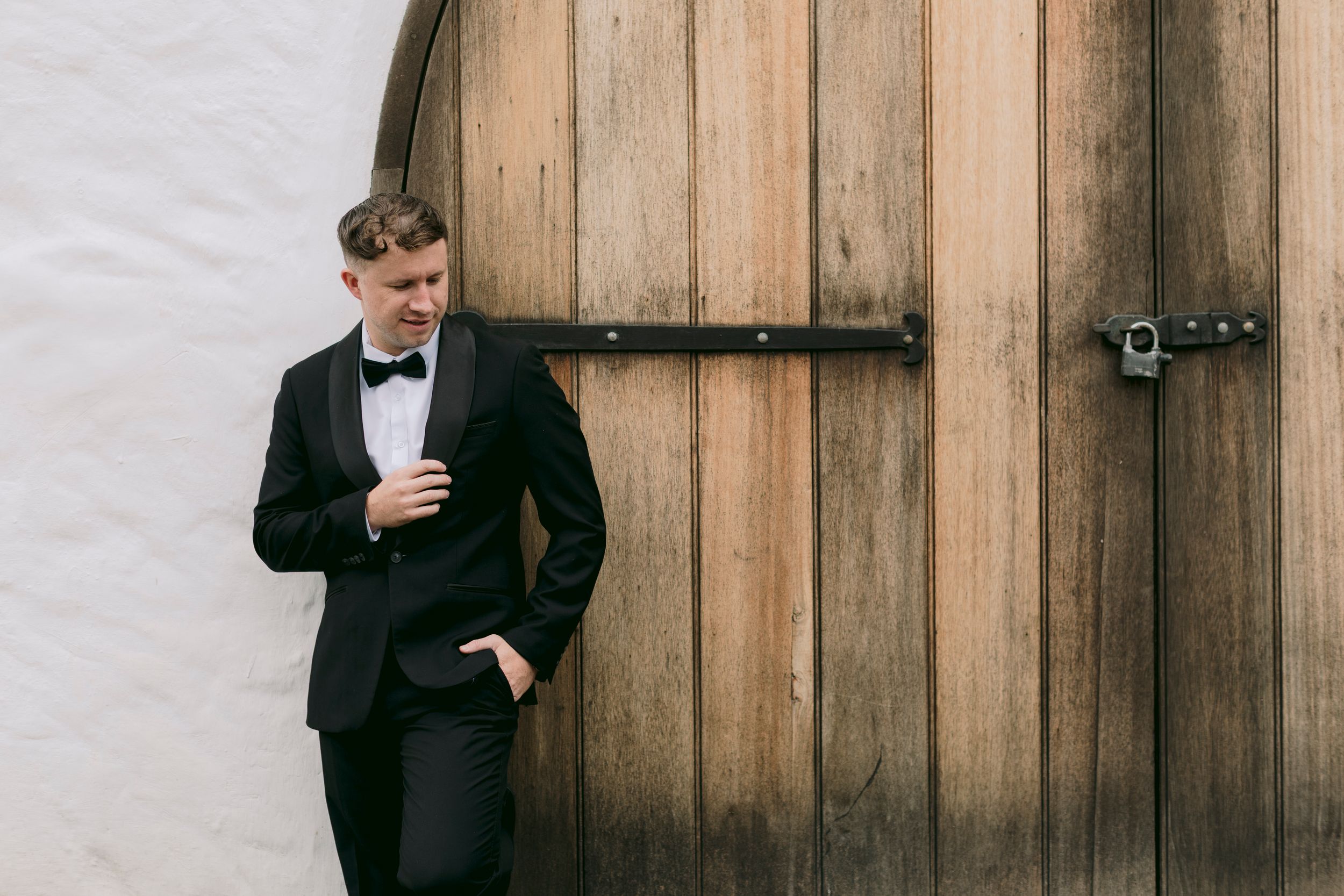 Groom in black tuxedo and bow tie leaning against rustic wooden barn door at wedding venue.