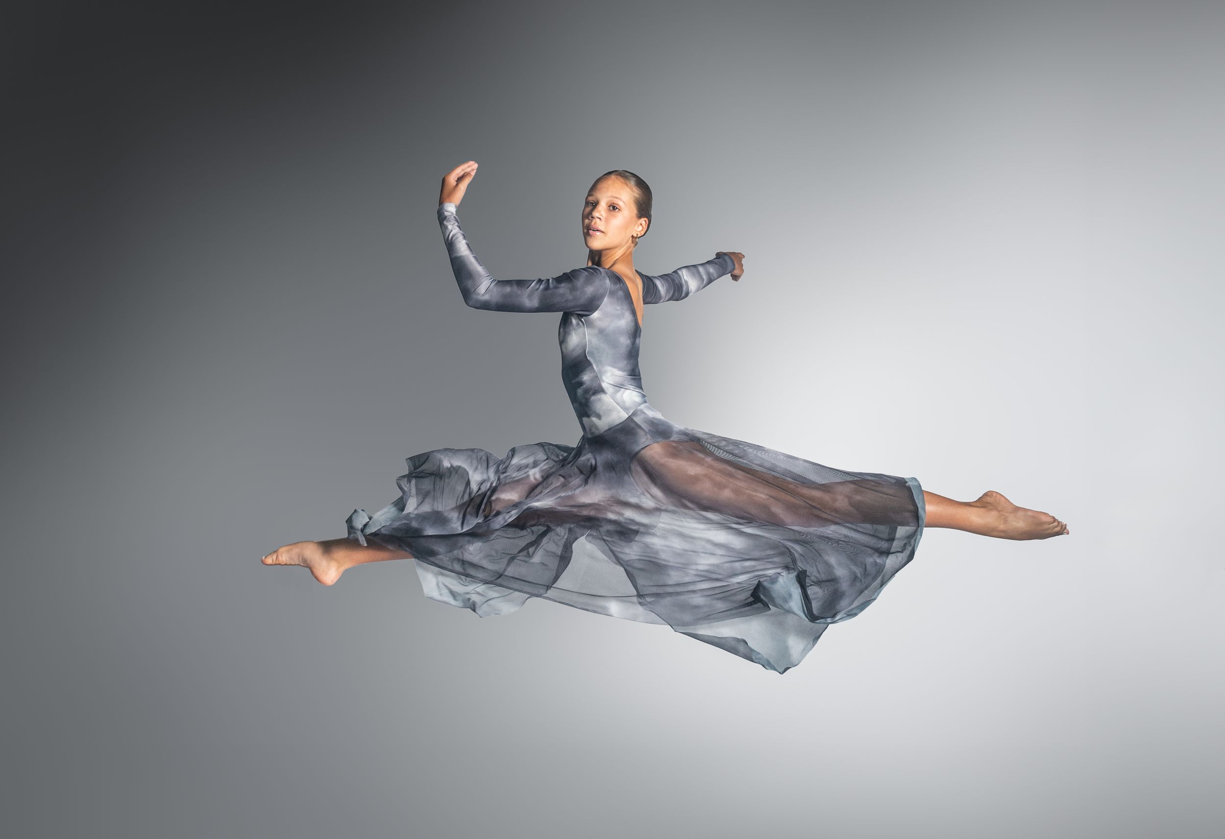 A graceful dancer leaps mid-air in a flowing gray dress, arms extended elegantly against a neutral studio background.