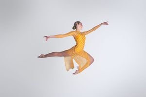 A dancer in a yellow dress leaps gracefully mid-air against a white background.