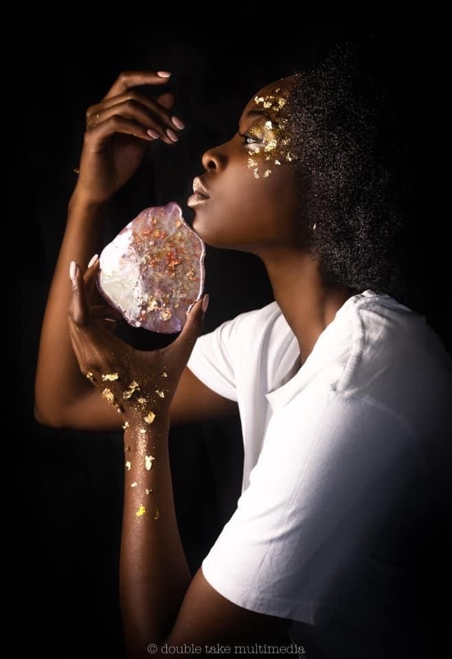 Woman holding glowing pink crystal with gold leaf accents on dark background, artistic portrait photography.