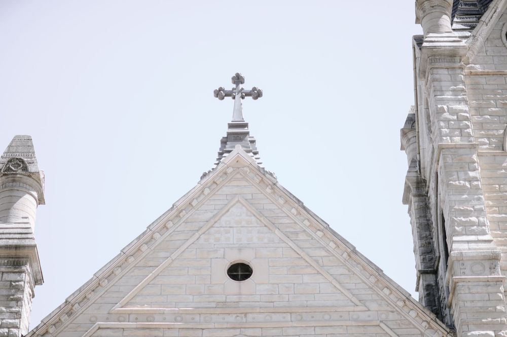White stone church facade with Gothic cross atop triangular gable against overcast sky.