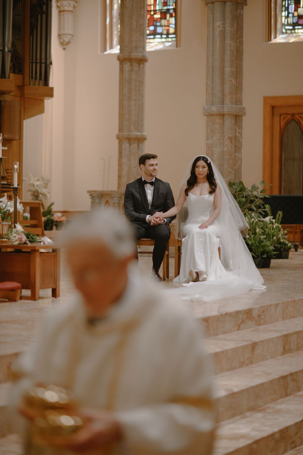 Bride and groom seated at church altar during wedding ceremony with priest in foreground.
