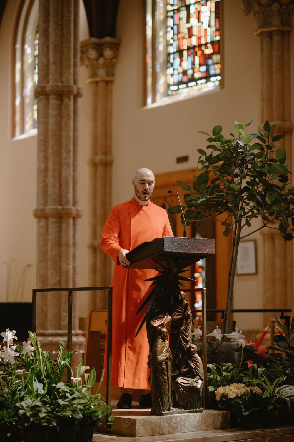 A monk in orange robes reads at a podium inside a church with stained glass windows and floral arrangements.