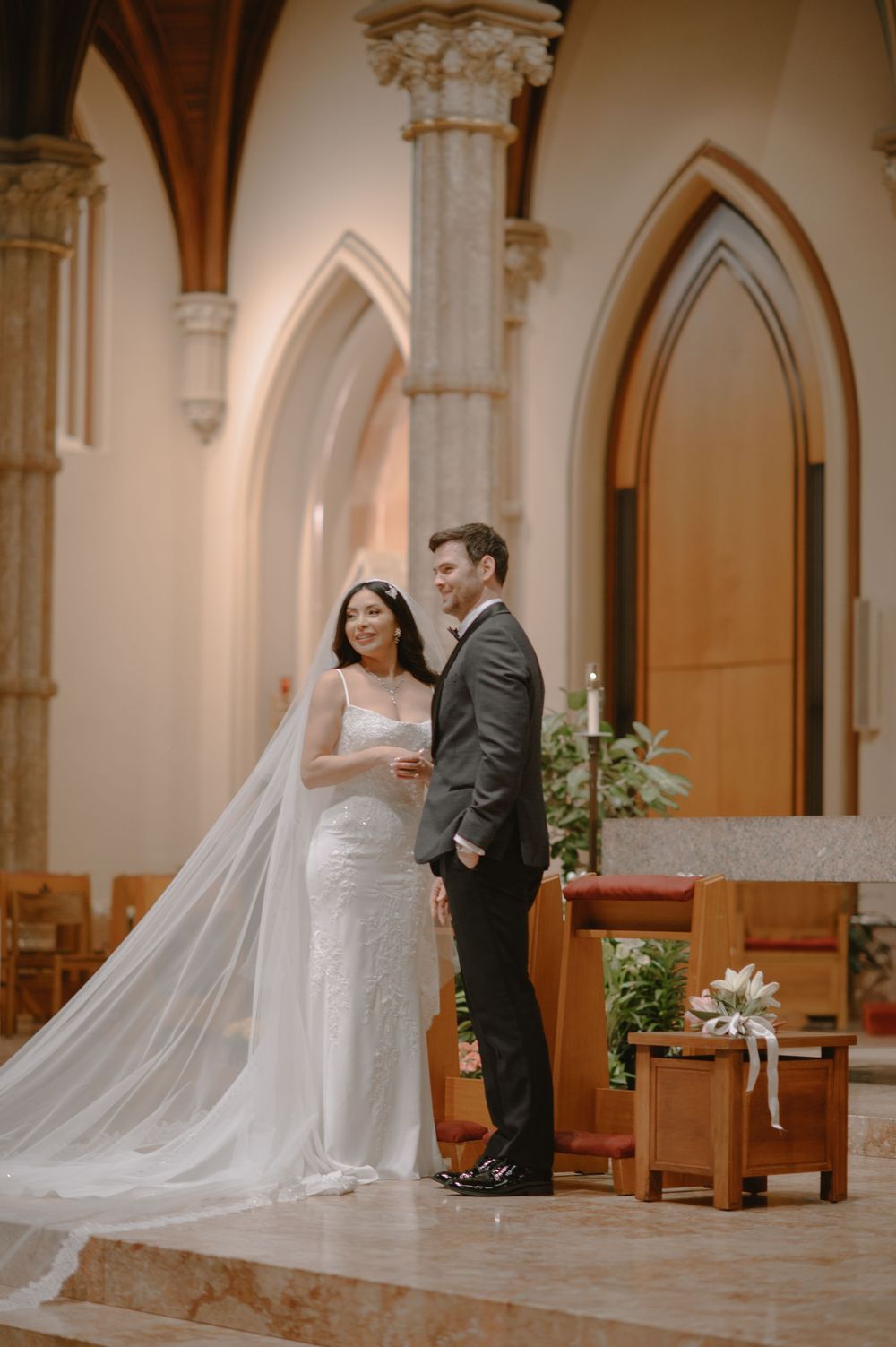 Bride and groom smiling together at church altar, bride wearing elegant gown with cathedral veil.