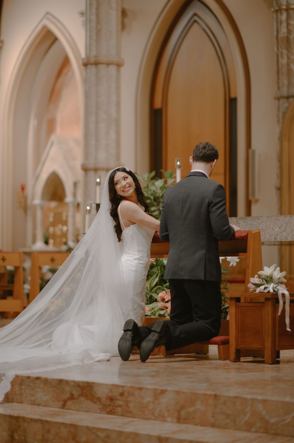 Bride smiling at altar during church wedding ceremony, wearing white gown and veil, kneeling beside groom in black suit.