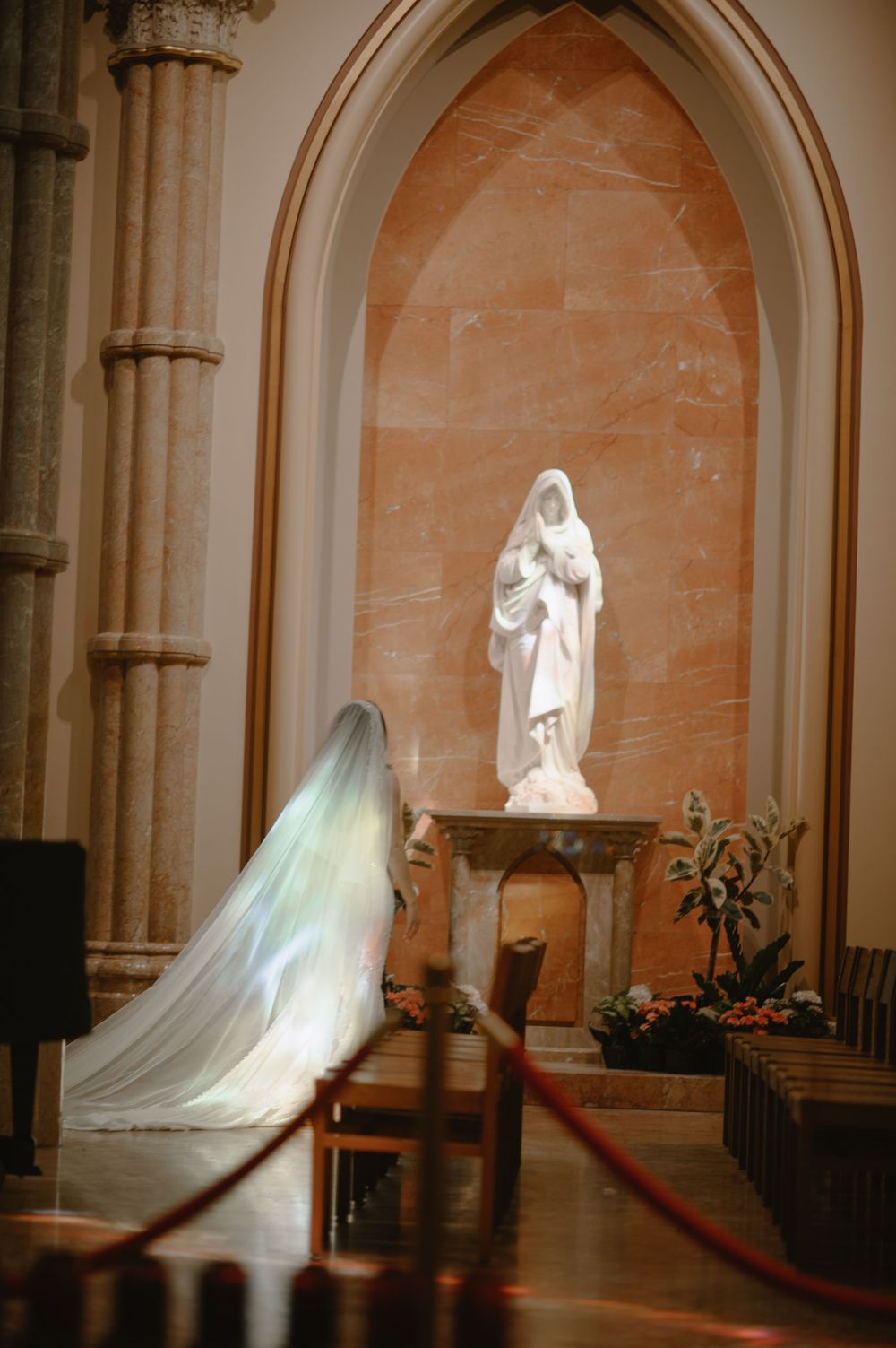 Bride in white gown and iridescent veil kneeling before a marble Virgin Mary statue in an elegant church.