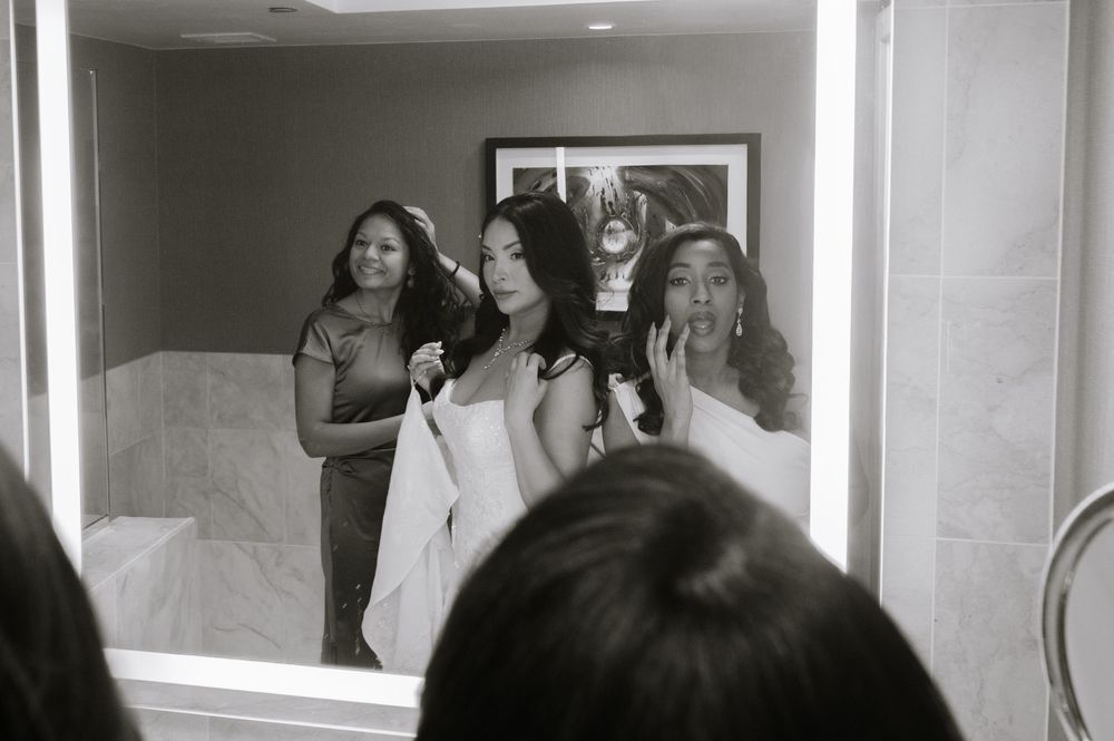 Black and white mirror reflection of three women getting ready, one having hair styled in elegant bathroom.