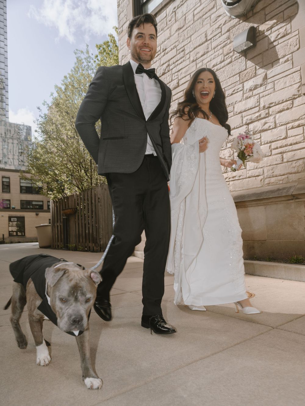 Couple in wedding attire with a gray pitbull dog posing outside a stone building on their wedding day.
