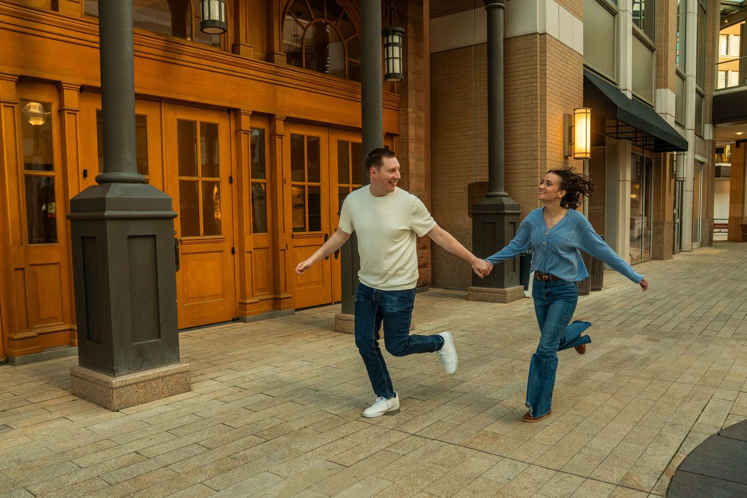 A joyful couple holds hands and runs together outside a building with warm wooden doors.