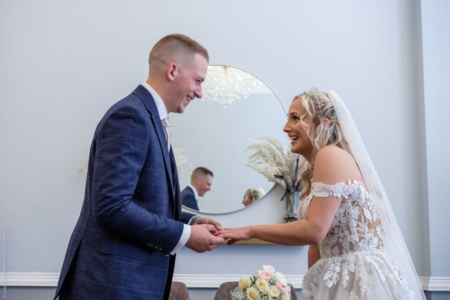 Bride and groom laughing together while exchanging rings at elegant wedding ceremony with mirror and floral decor.