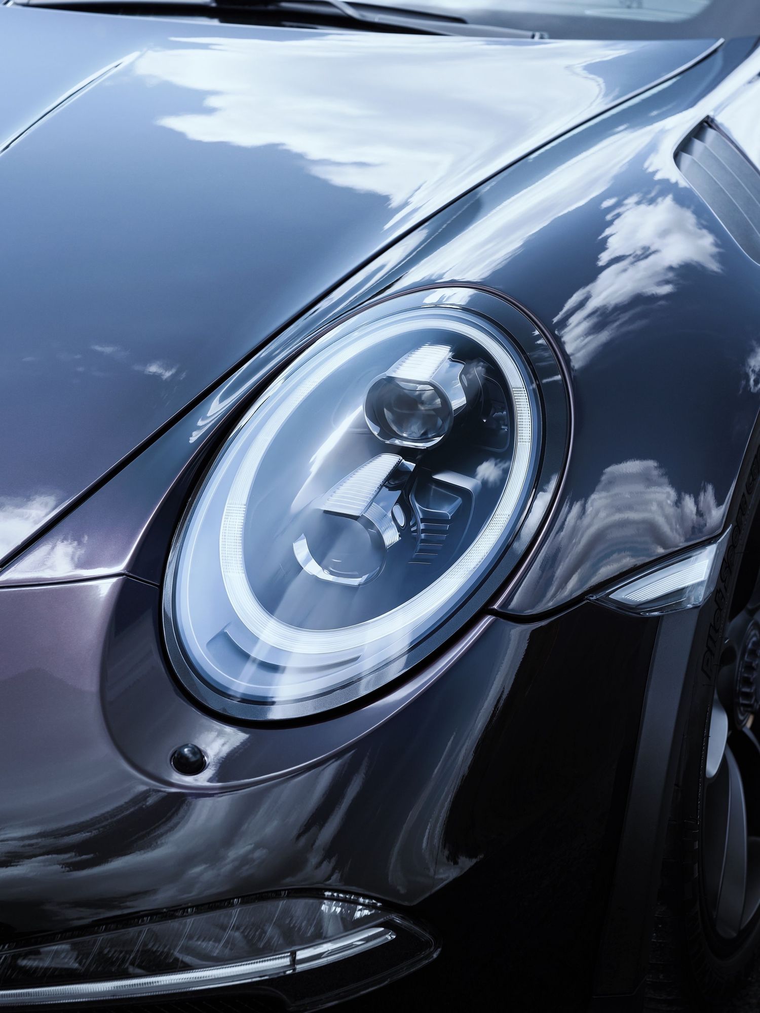 Close-up of a sleek Porsche 911 headlight with LED ring, reflecting clouds on its glossy dark gray hood.