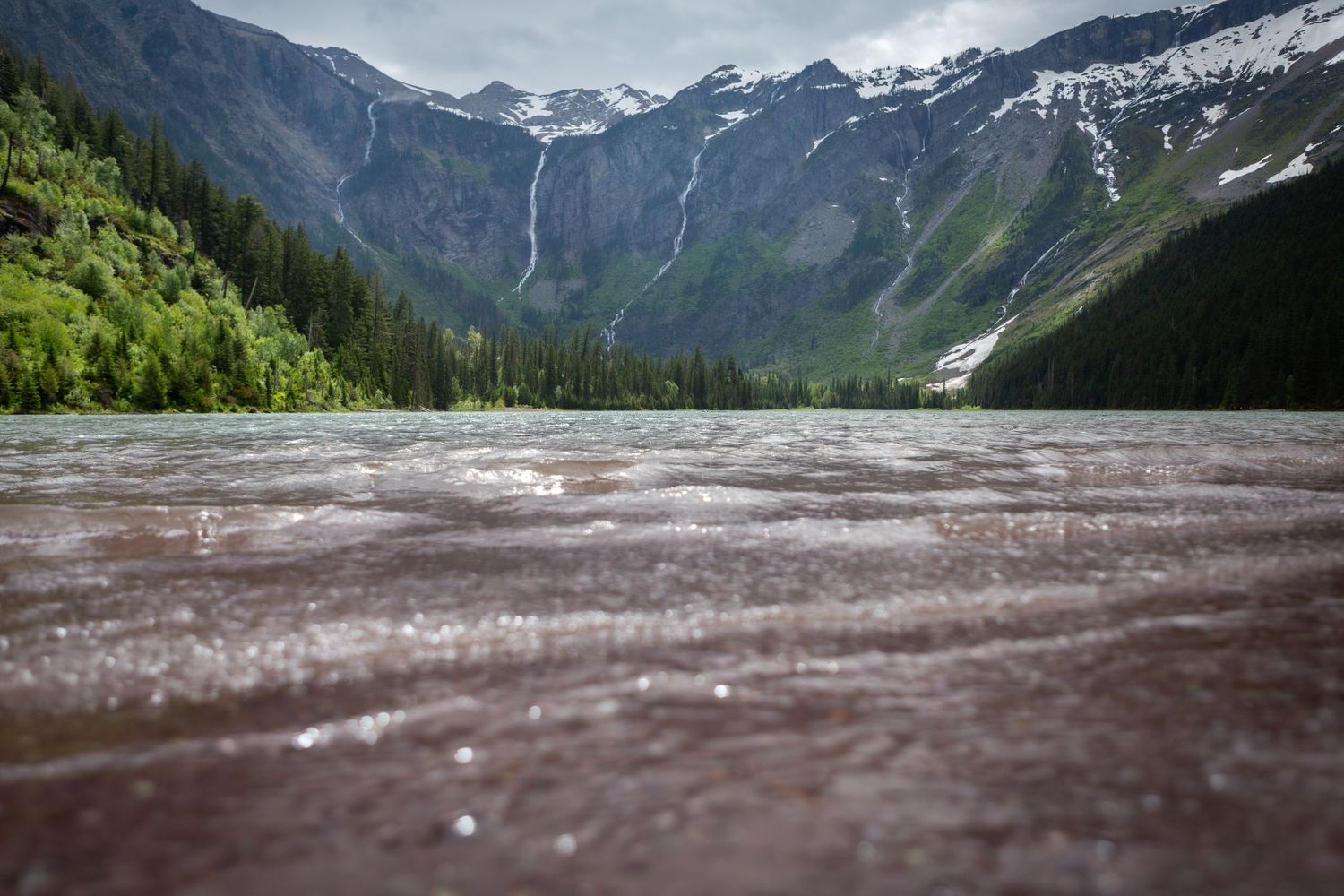 Rain falling on a mountain lake with snow-capped peaks, evergreen forests, and a scenic valley in the background.