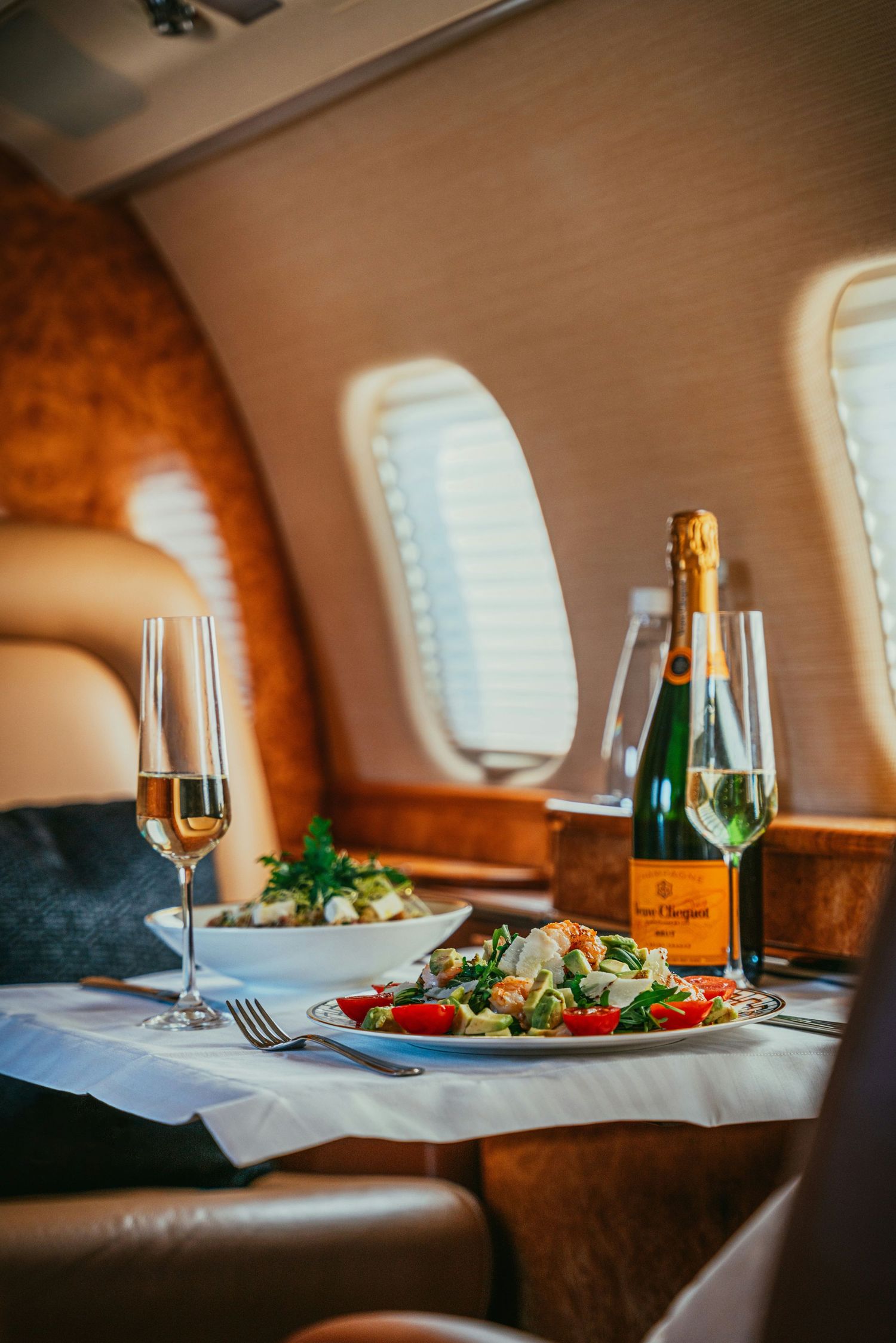 Elegant dining setup on a private jet with Veuve Clicquot champagne, fresh salads, and crystal glasses by oval windows.