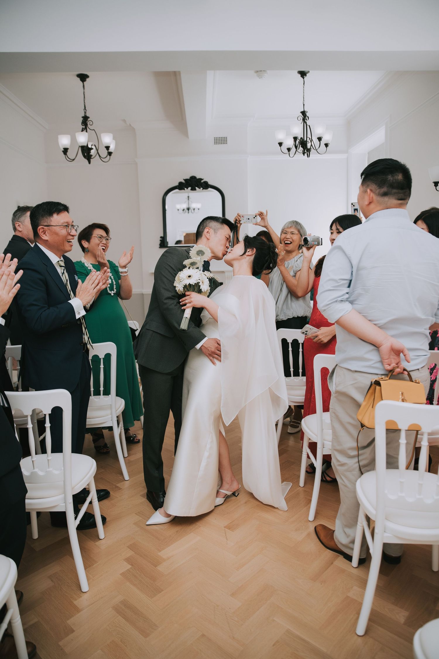 Joyful newlywed Asian couple celebrating after wedding ceremony, bride raising bouquet, guests applauding in elegant white venue.