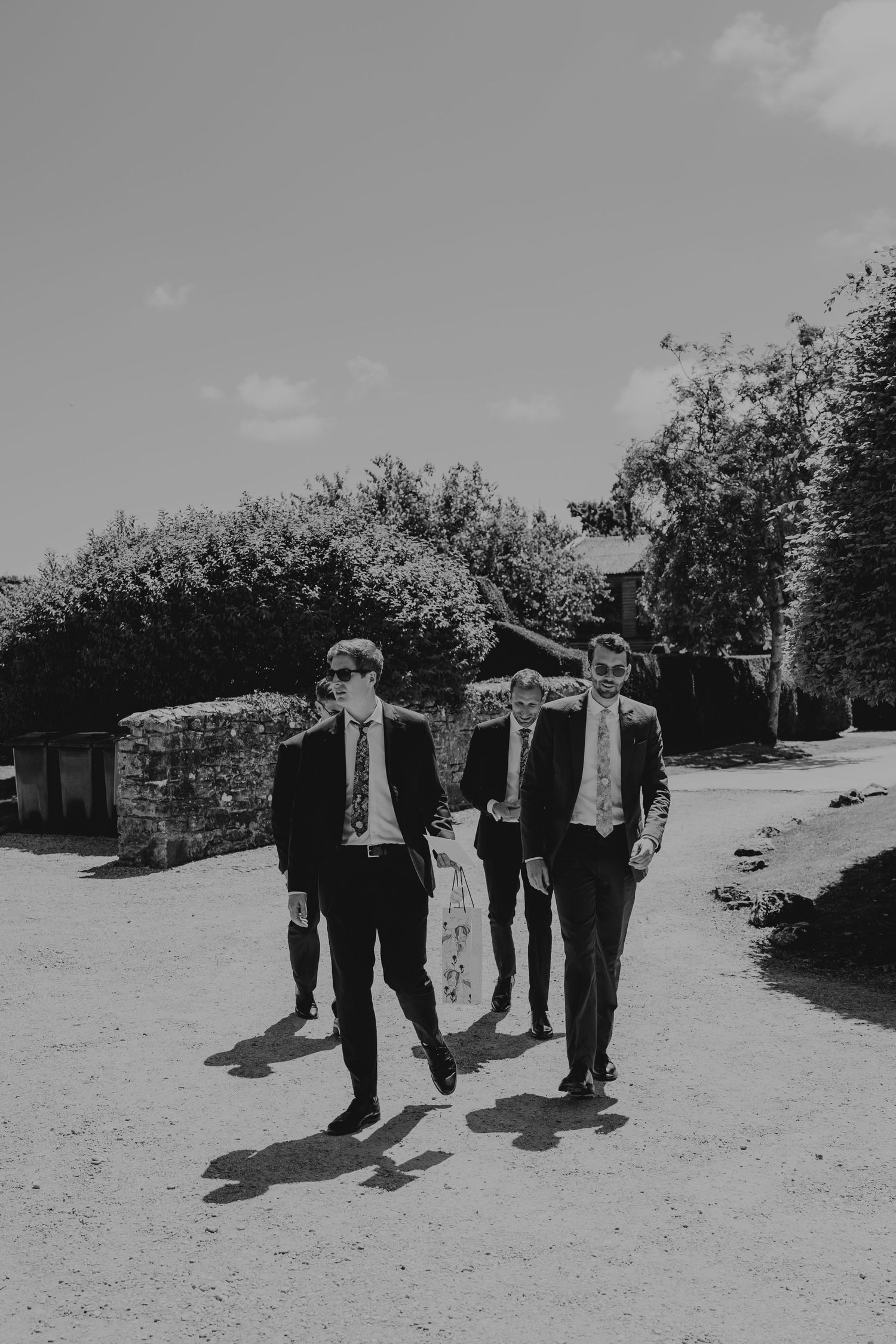 Three groomsmen in suits and ties walking confidently down a gravel path outdoors in a black and white wedding photo at Kingston Estate.