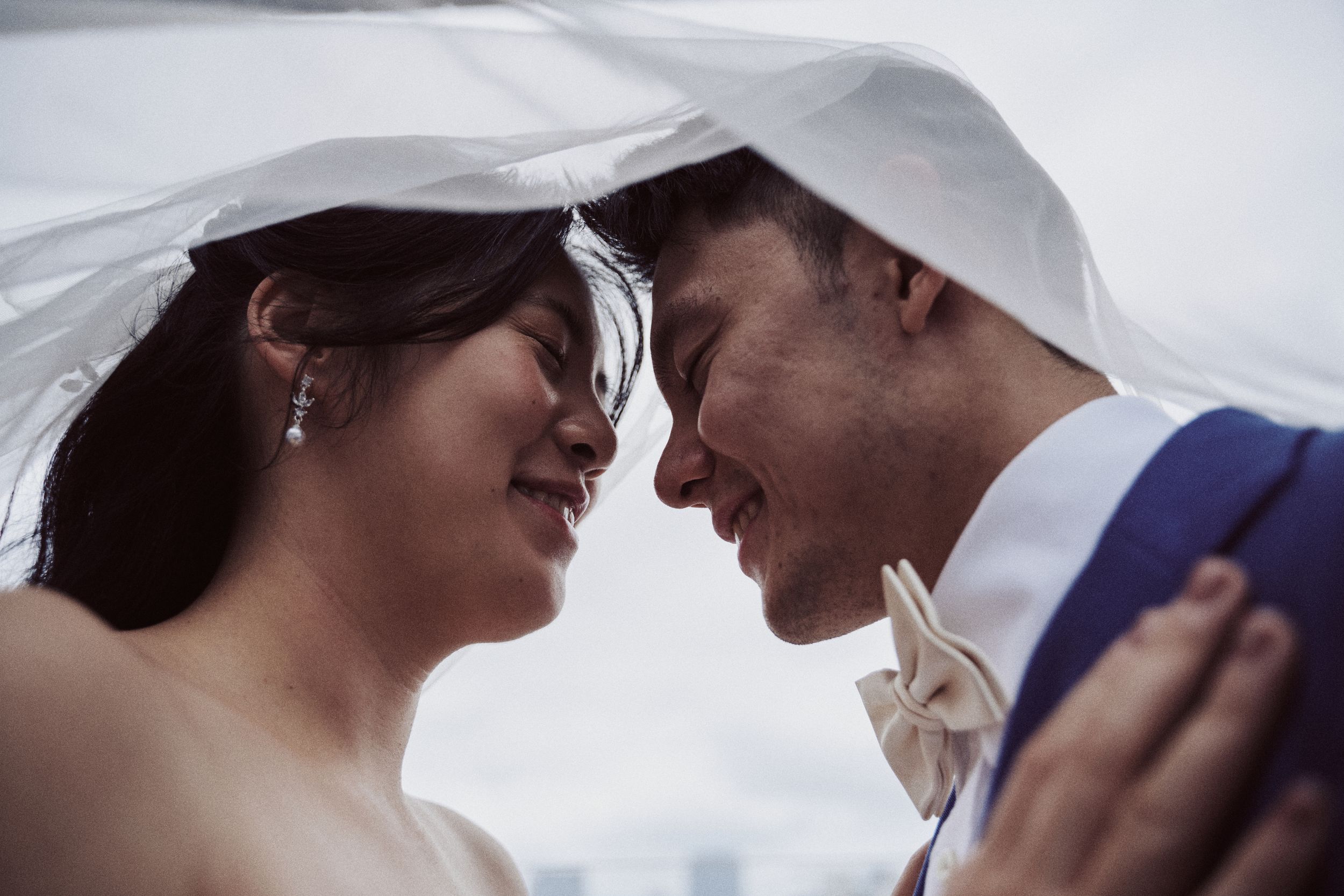 A bride and groom share an intimate moment under a wedding veil, smiling at each other in a romantic close-up.