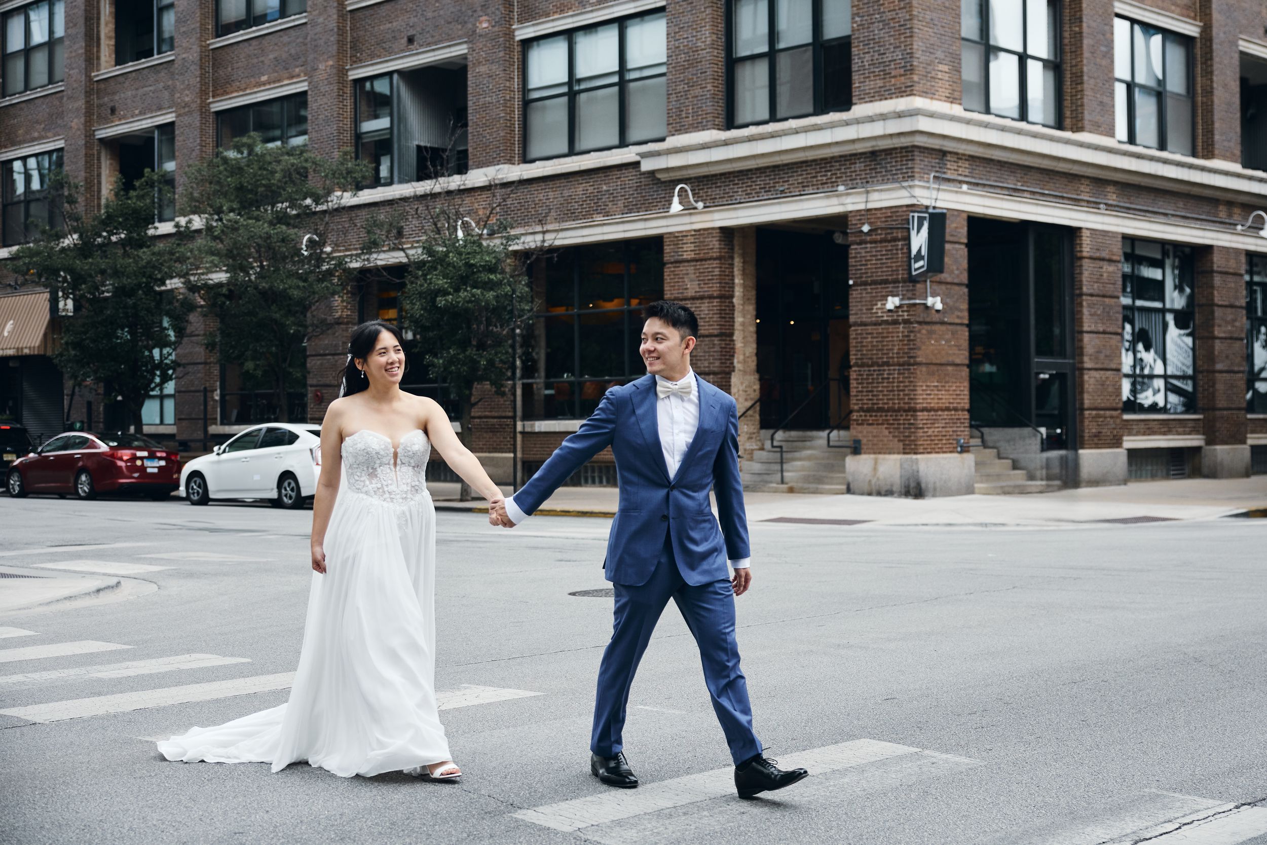 Bride and groom holding hands crossing a city street, bride in white gown, groom in blue suit, urban brick building backdrop.