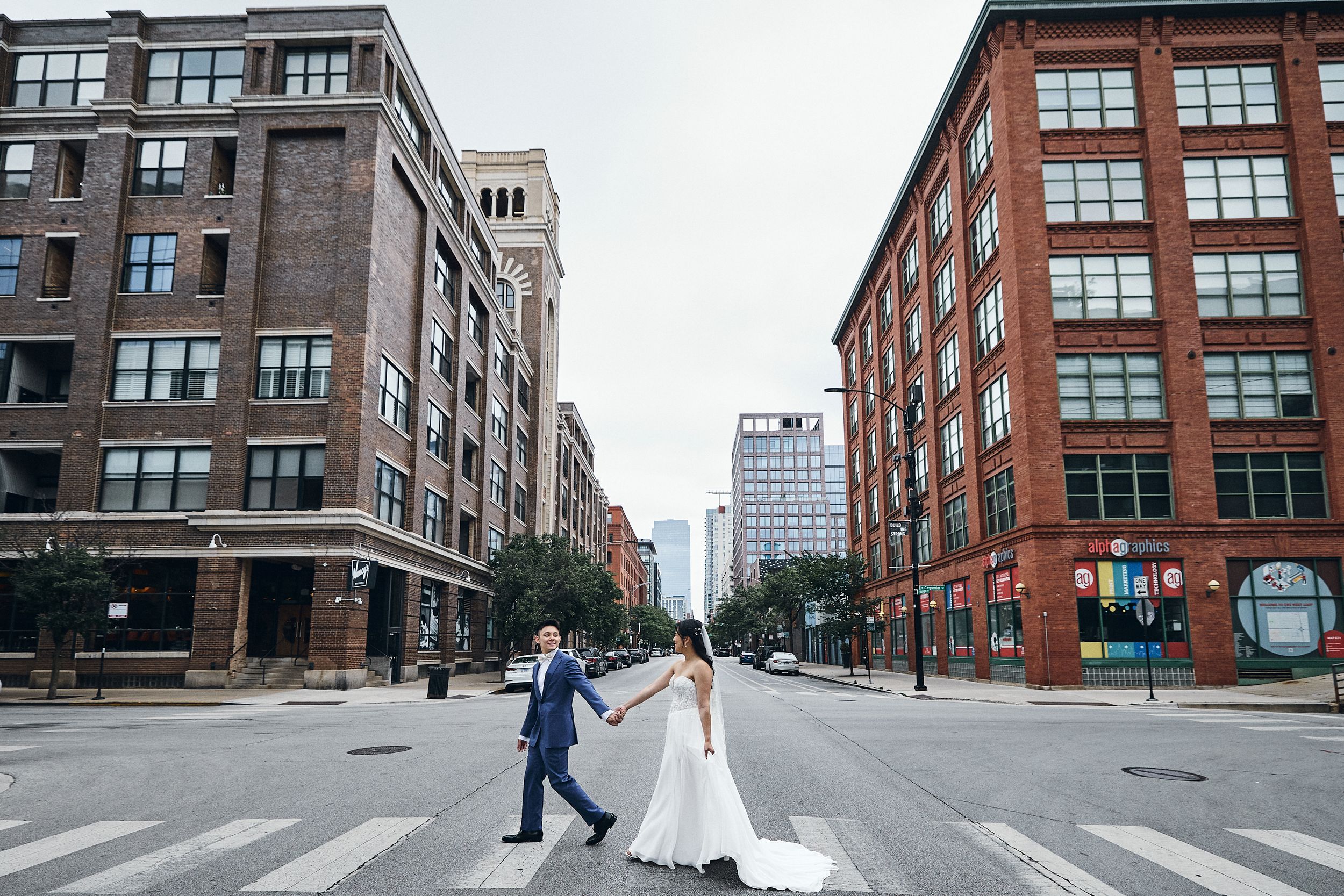 Bride and groom crossing a city street intersection in urban downtown, wearing wedding attire, surrounded by brick buildings.