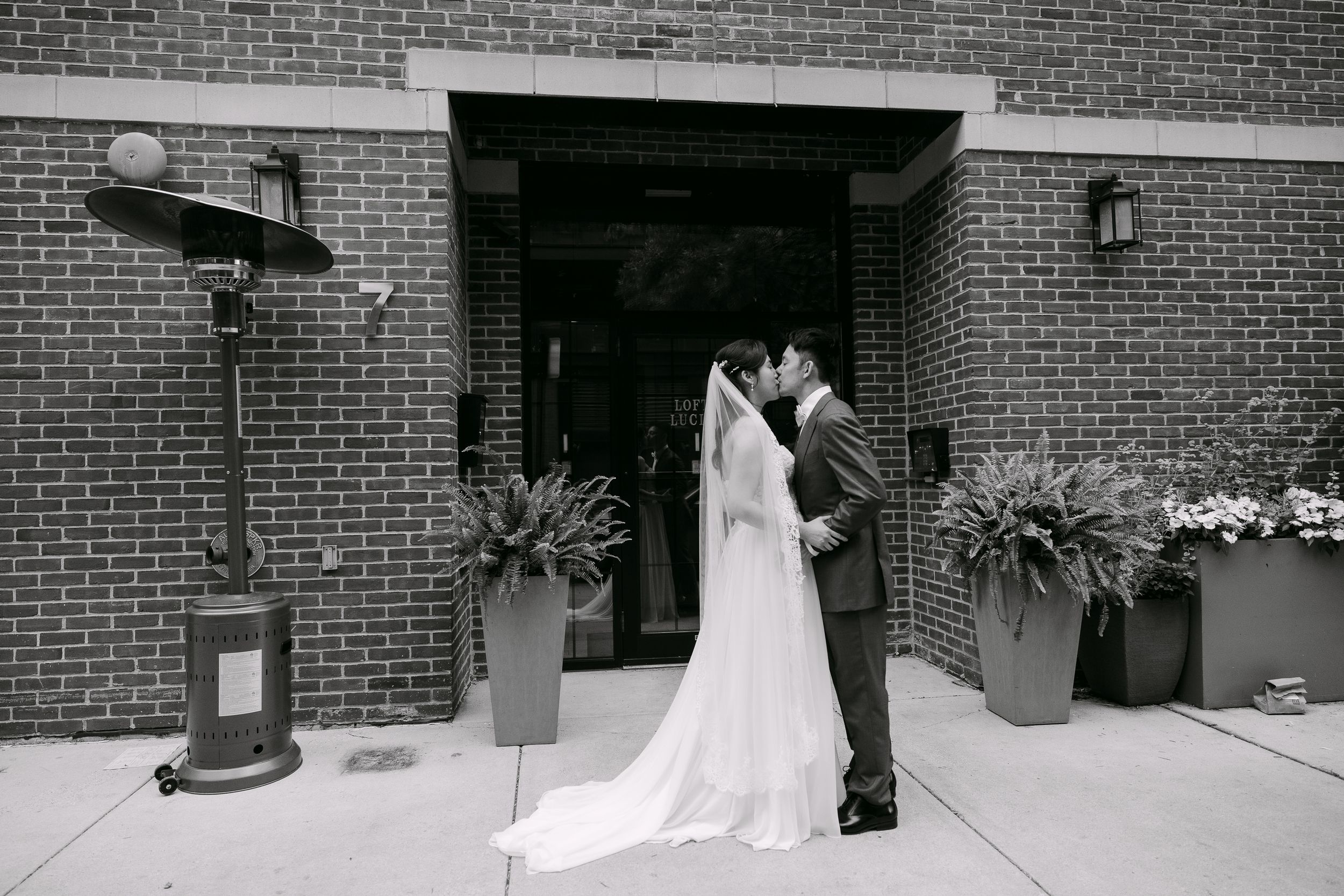 Black and white photo of a bride and groom sharing a kiss outside a brick building on their wedding day.