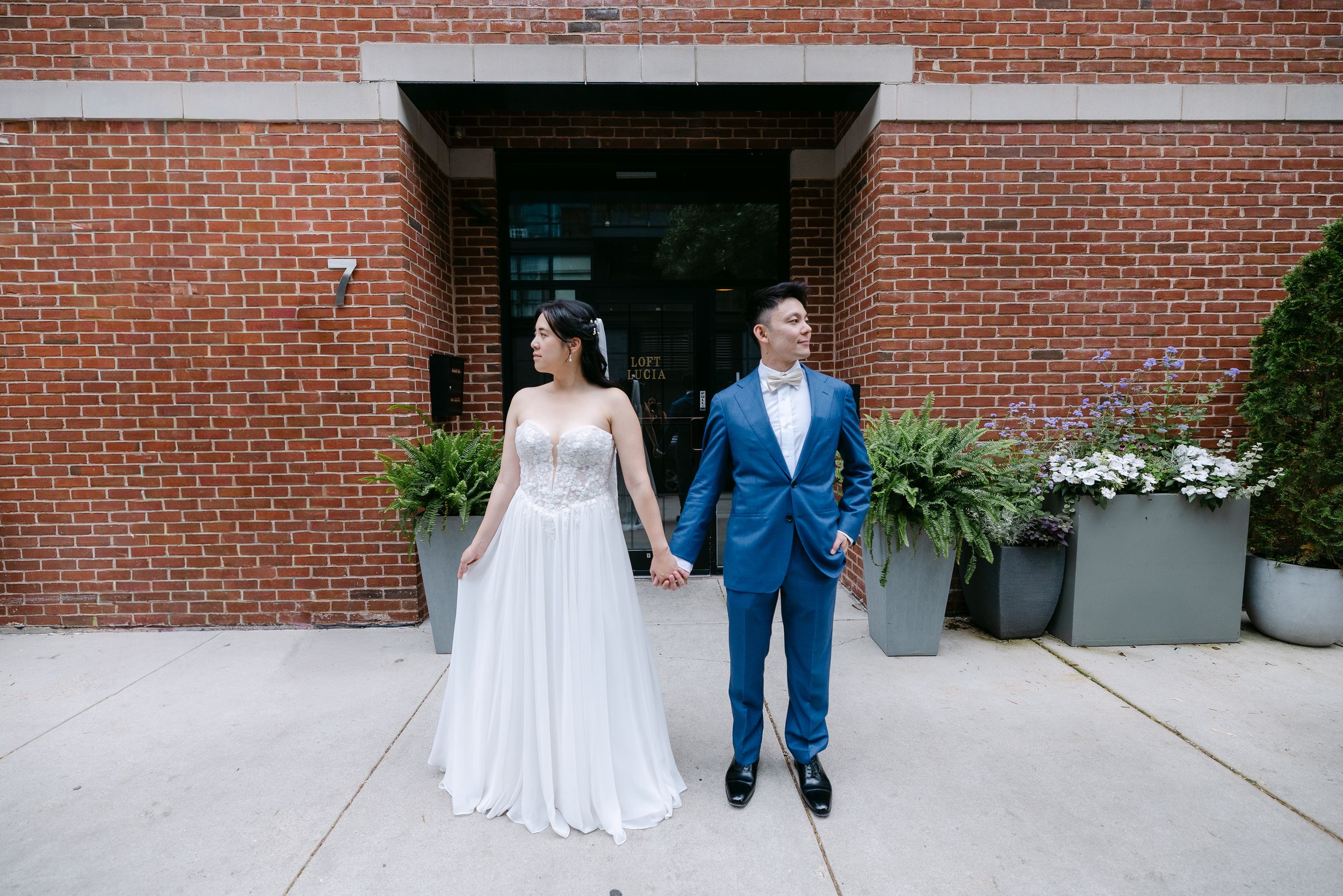 Asian couple in wedding attire holding hands outside brick building, bride in white gown, groom in blue suit with bow tie.