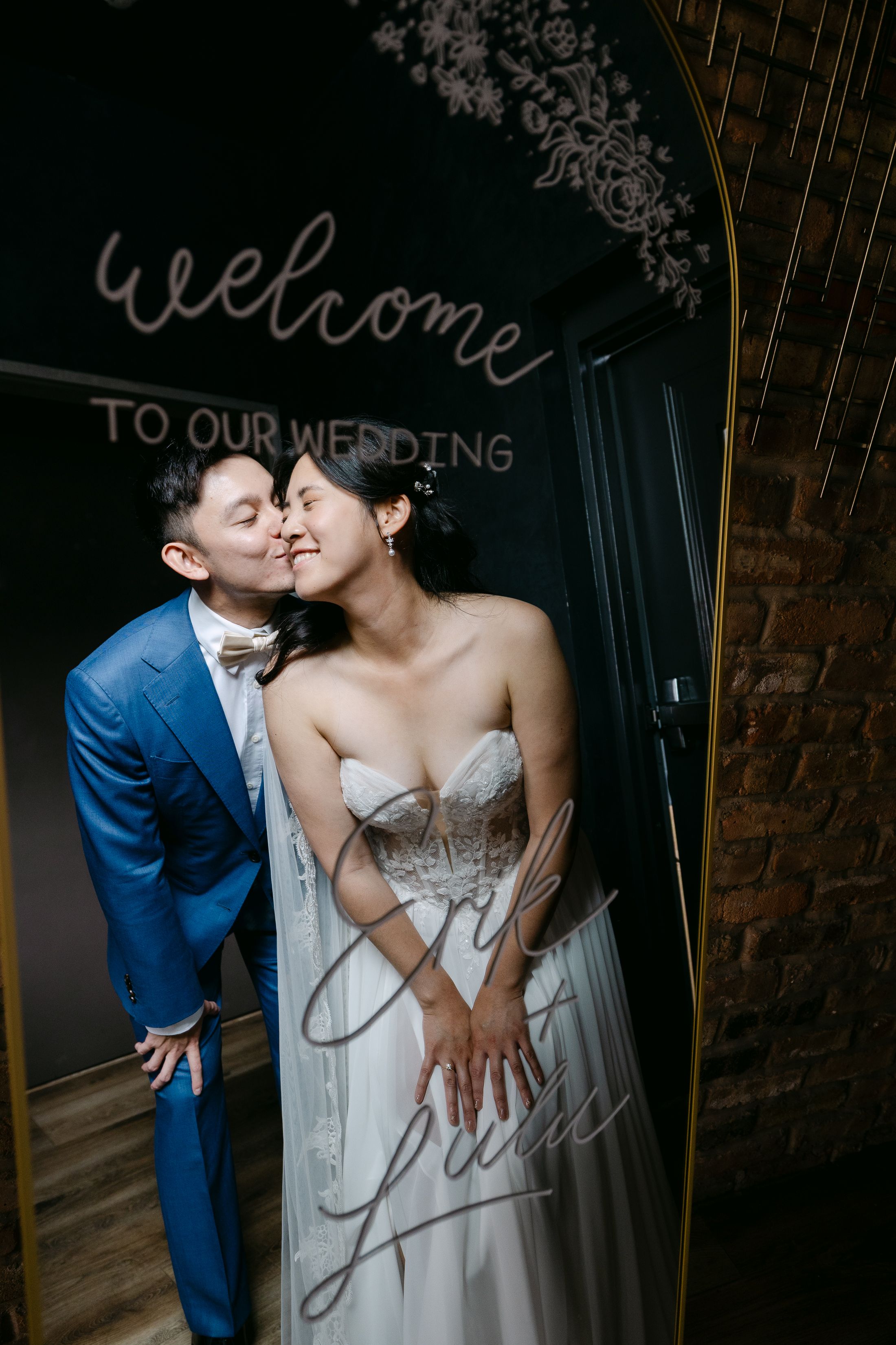 Groom in blue suit kissing bride in strapless gown beside a welcome to our wedding sign.