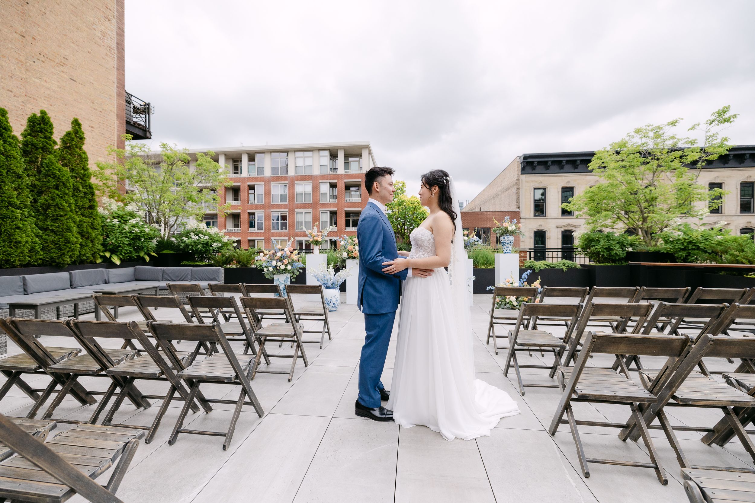 Bride and groom share a romantic moment on a rooftop wedding venue with wooden chairs and city buildings backdrop.