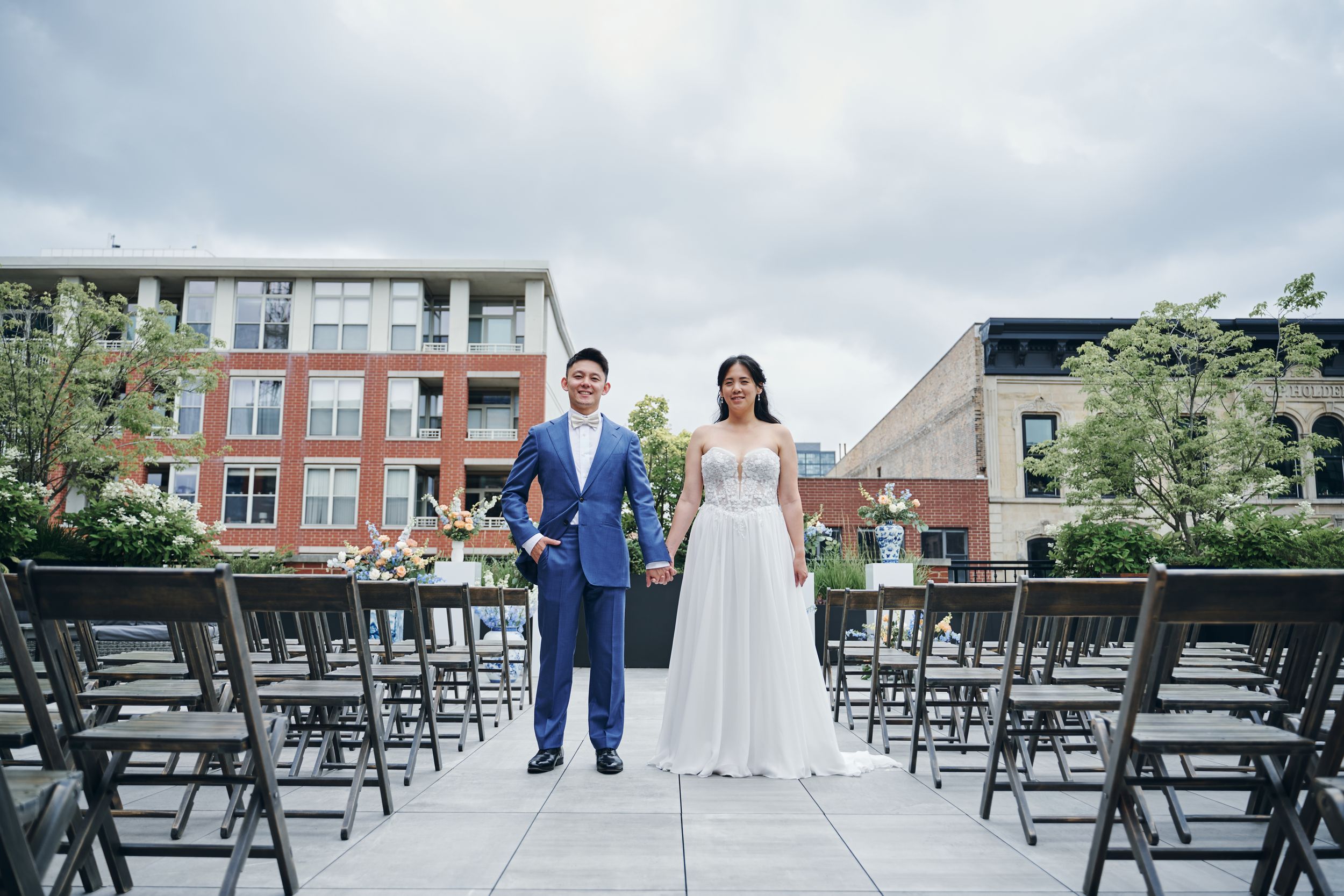 Bride and groom holding hands at outdoor wedding ceremony venue with rows of chairs and urban buildings in background.