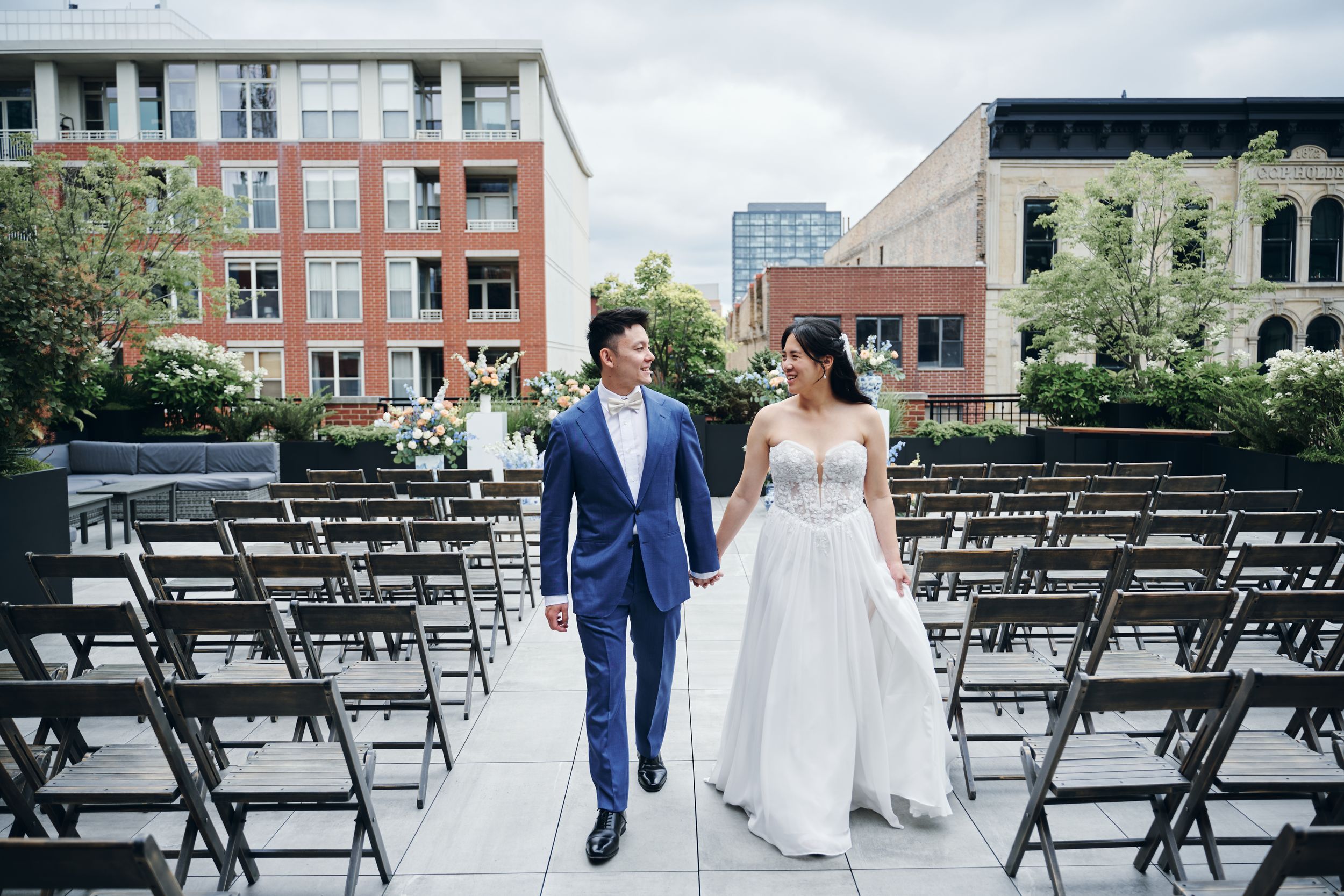 Bride and groom walk hand-in-hand through outdoor wedding venue with rows of black chairs and urban buildings backdrop.