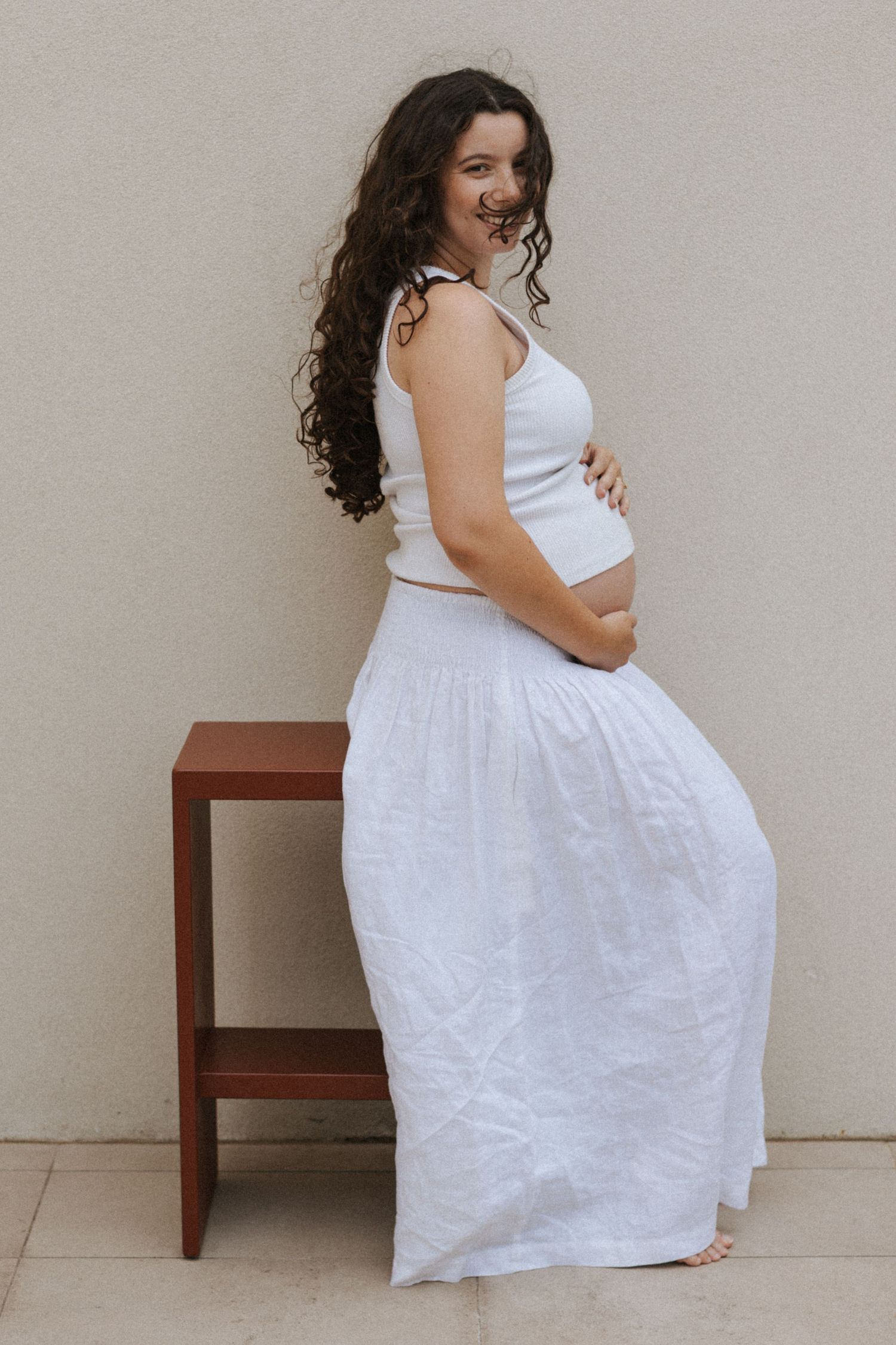 Pregnant woman in white crop top and maxi skirt cradling baby bump, smiling beside wooden stool outdoors.