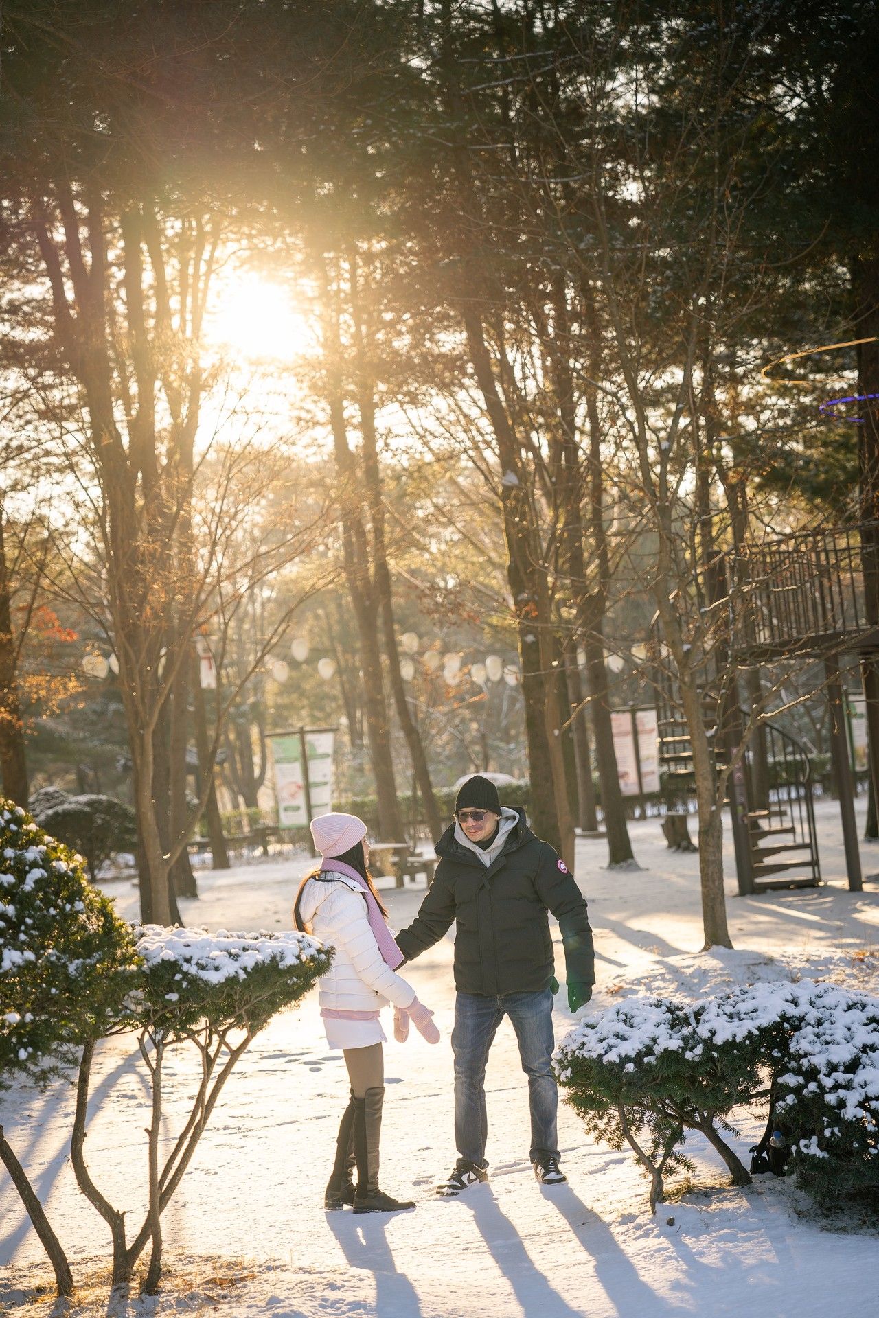Couple holding hands in snowy winter park at golden sunset, surrounded by snow-covered trees and bushes.