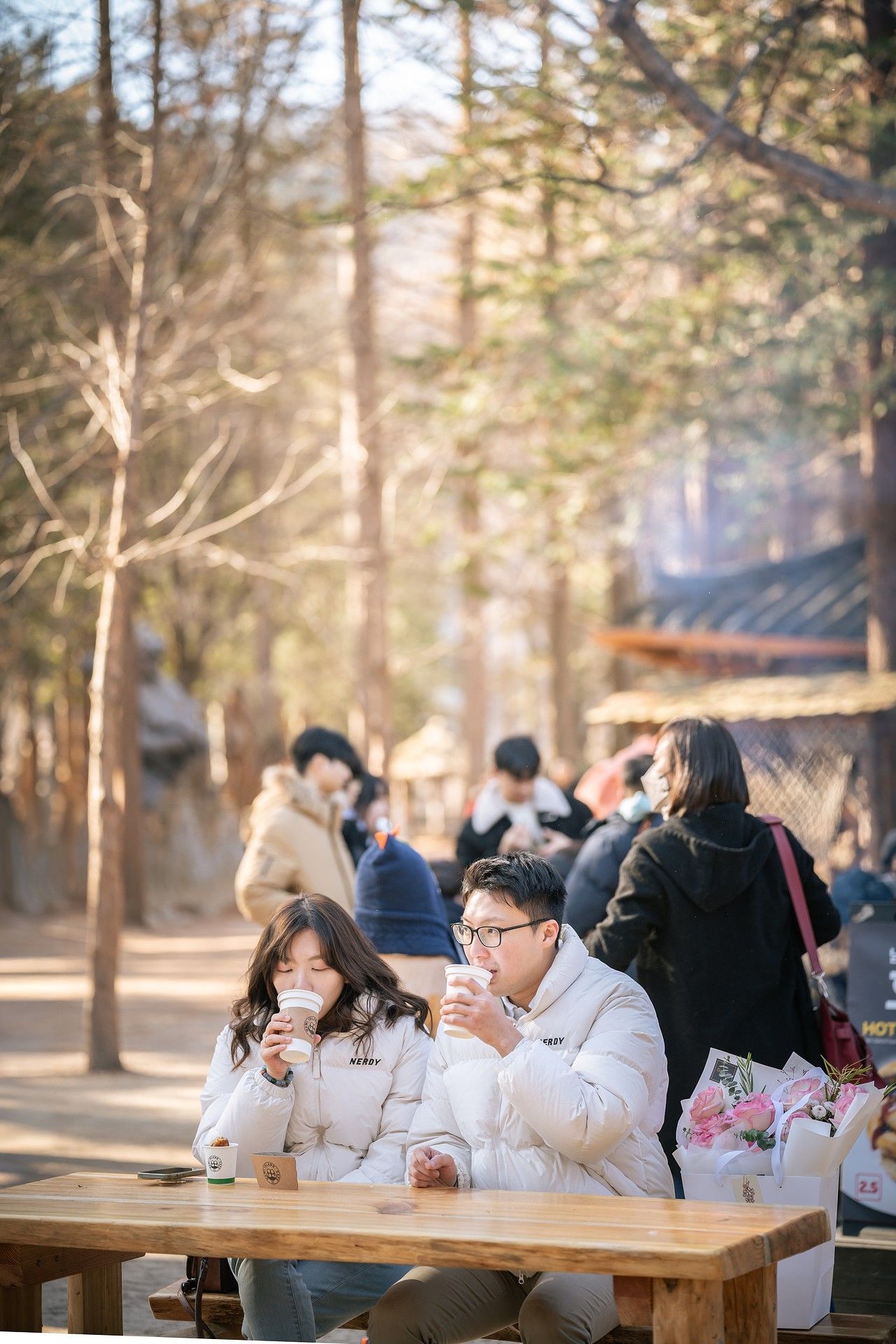 Couple drinking hot beverages at outdoor wooden table in sunlit forest park during winter.