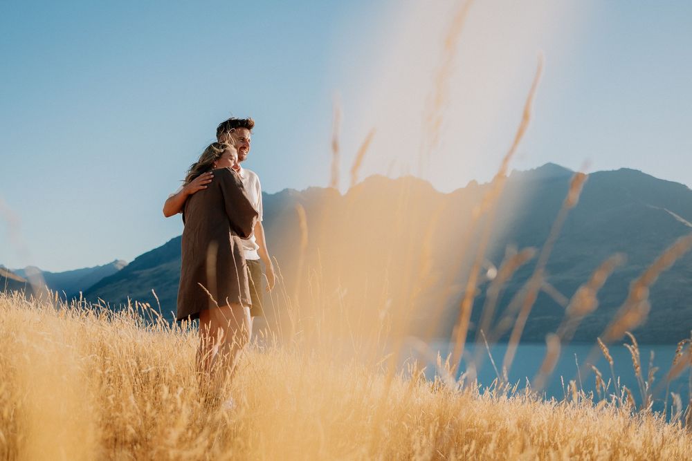 A couple walks through a golden wheat field at sunset with mountains in the background.