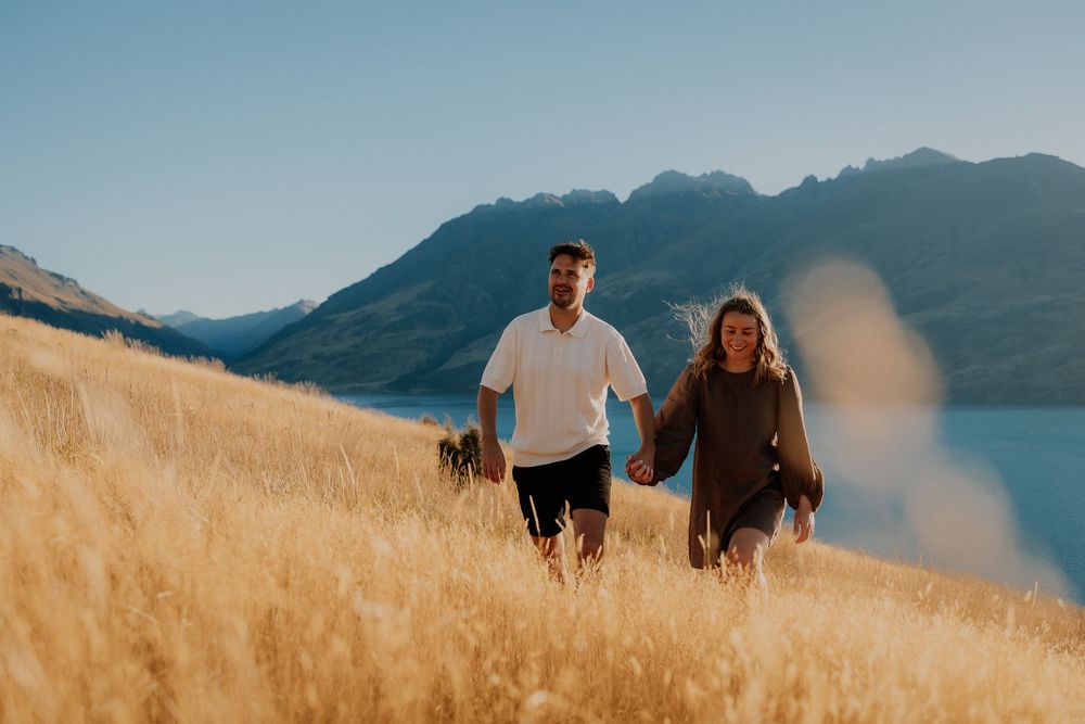Two people walk through golden grass on a hillside overlooking a mountain lake on a sunny day.