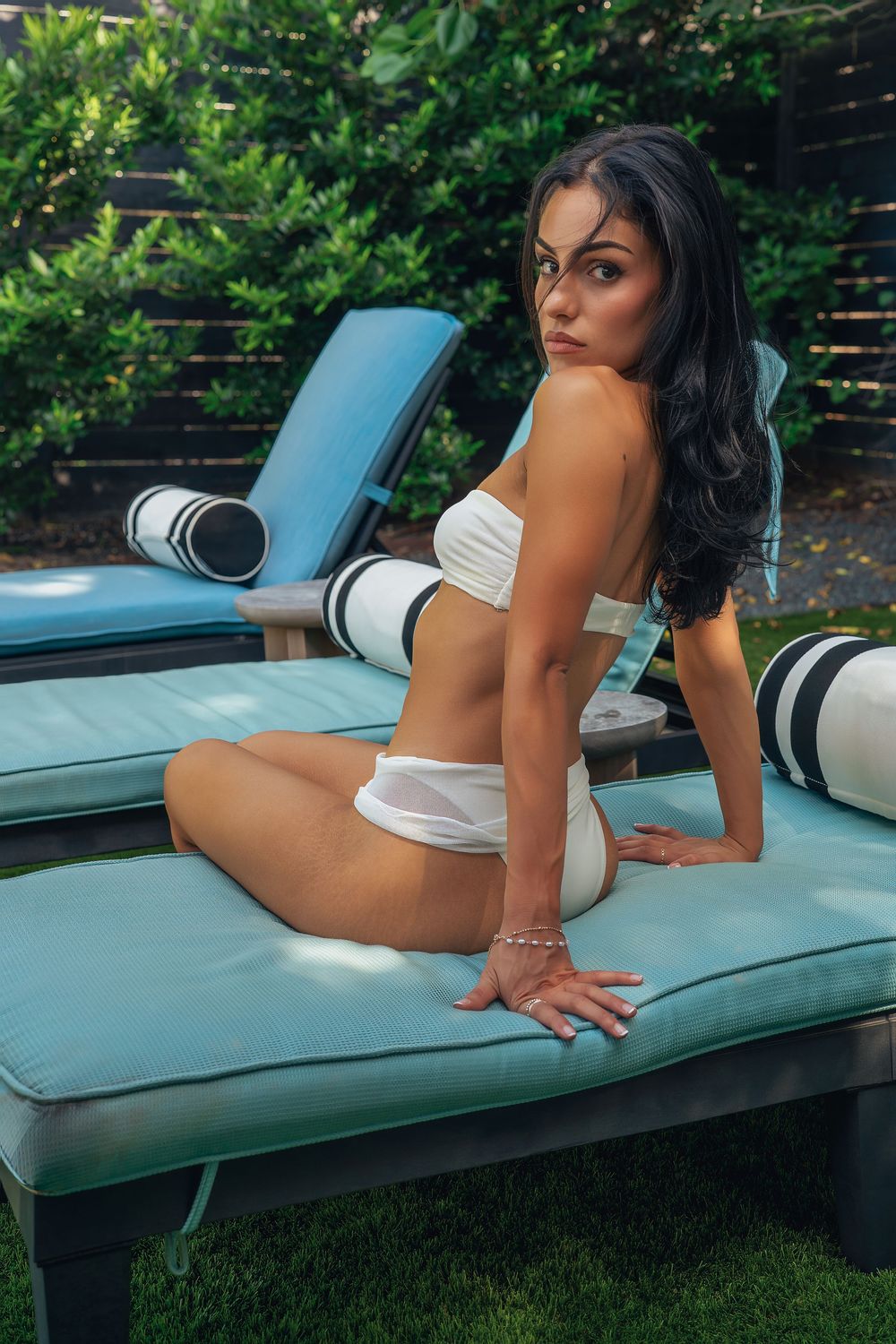 Woman in white bikini posing on blue lounge chair outdoors near lush green hedges.