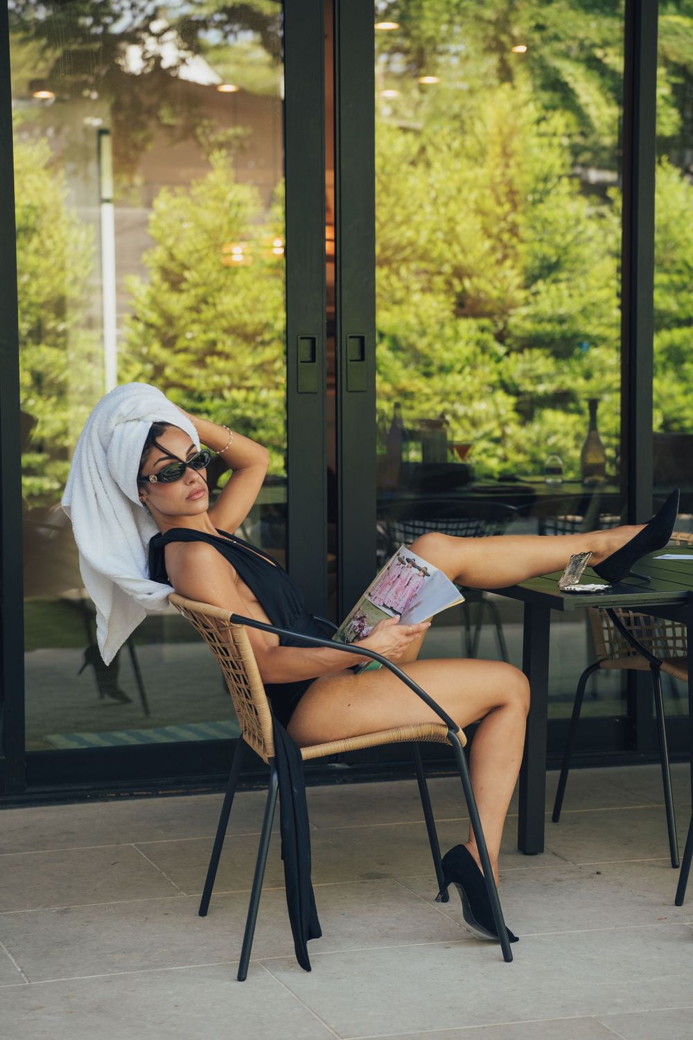 Woman in black swimsuit and towel turban relaxes on patio chair with magazine and heels.