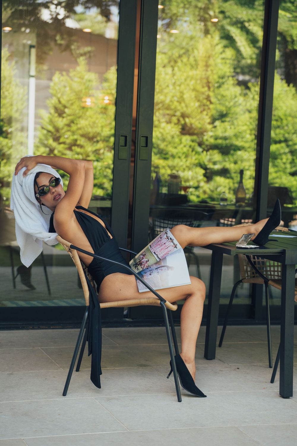 Woman relaxing on chair outdoors, wearing sunglasses and towel on head, holding magazine, with lush green trees in background.