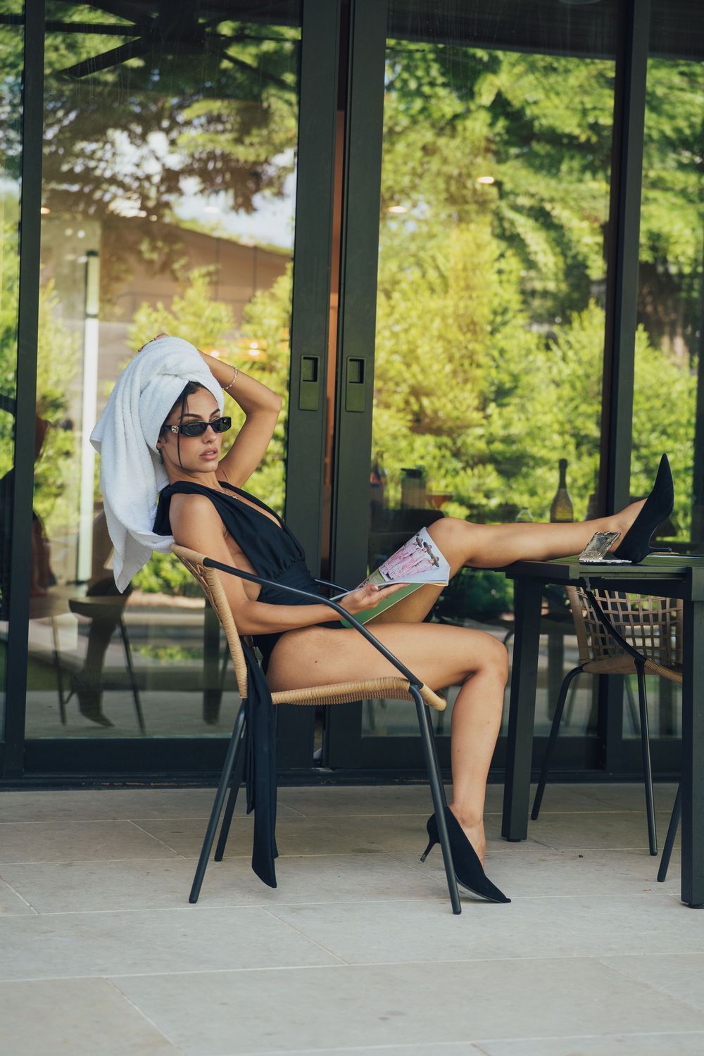 Woman in black top, denim shorts, heels, and towel turban relaxing on patio chair with sunglasses outdoors.