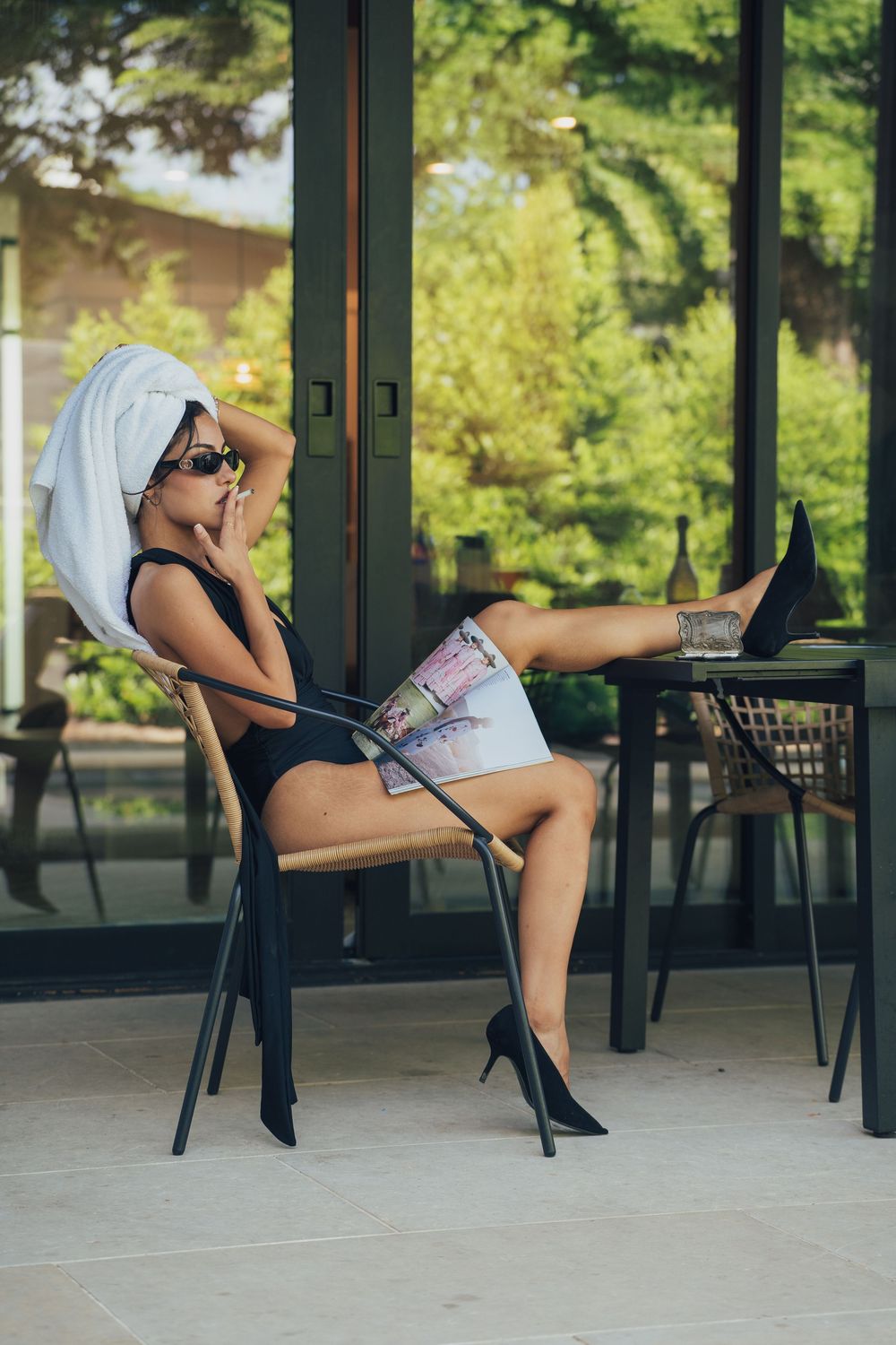 Woman relaxing on patio chair in black bodysuit, shorts, heels, and towel turban, with laptop on table outdoors.