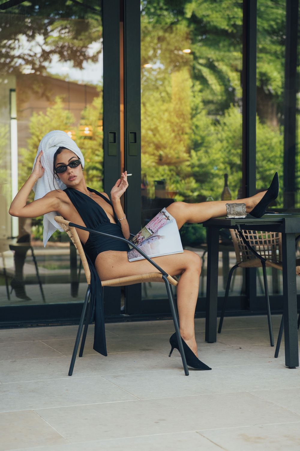 Stylish woman in black swimsuit and towel turban poses on patio chair with cigarette and heels.