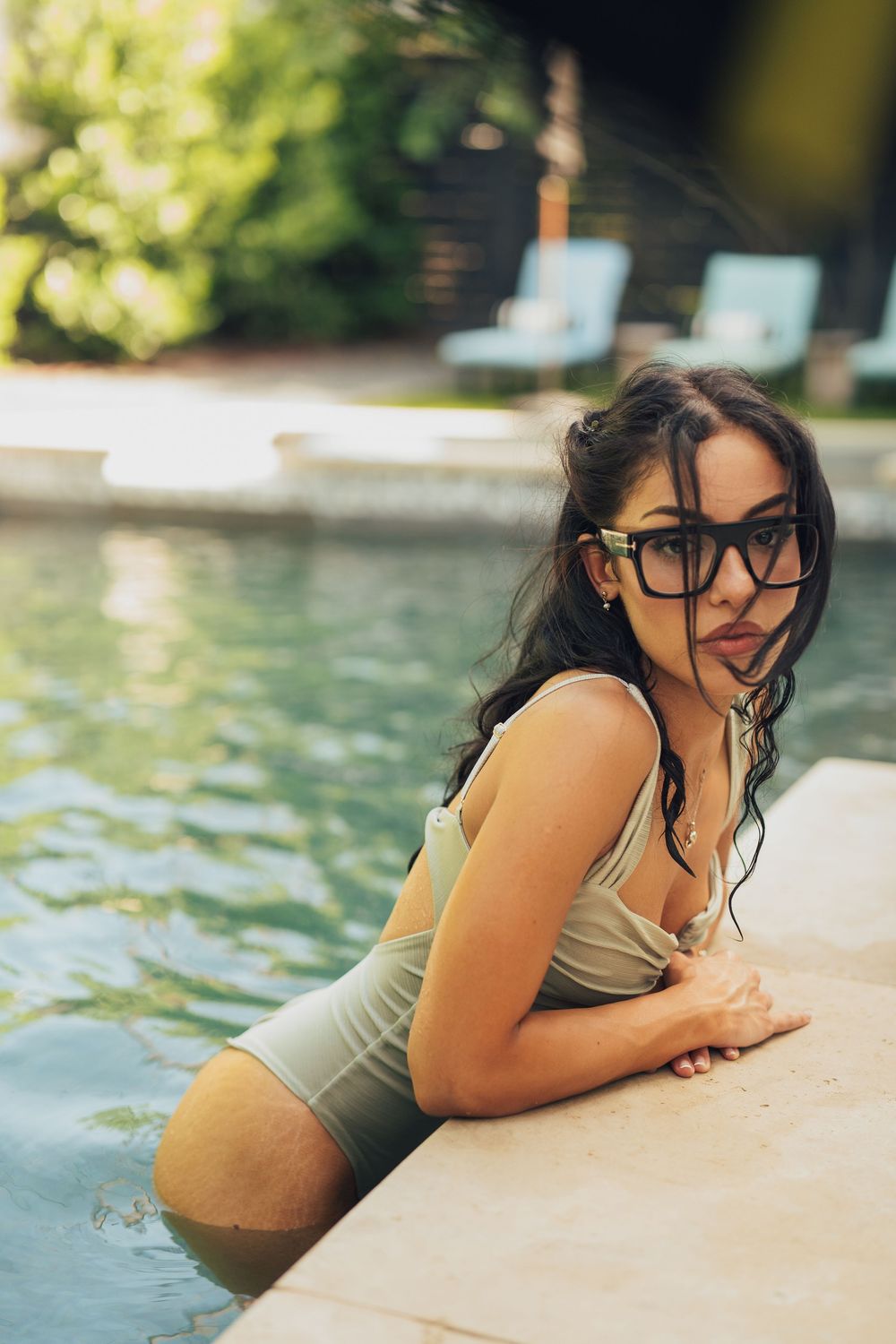 Young woman in olive swimsuit and glasses leans on pool edge with wet hair.