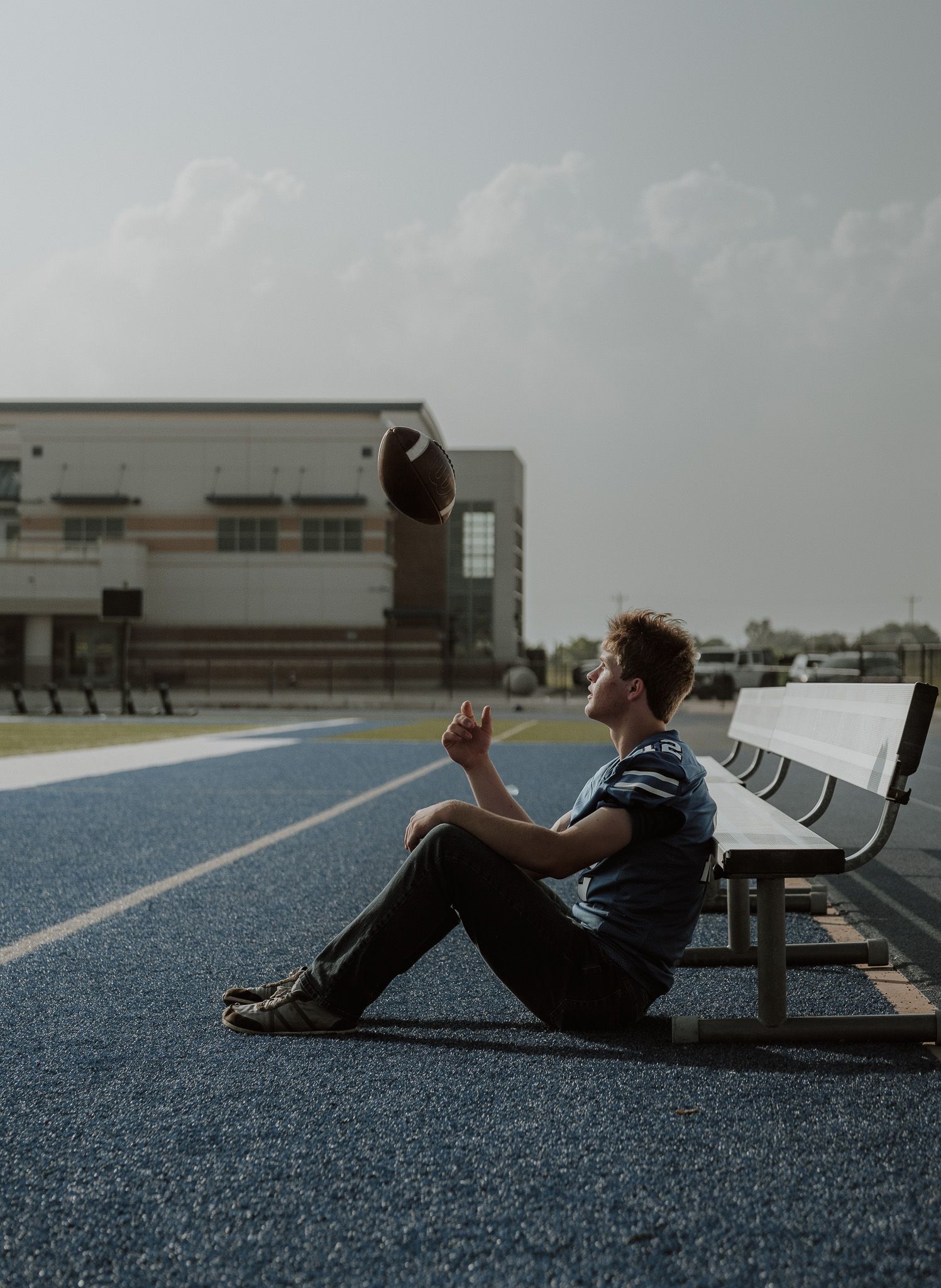 A young man sits on a blue track tossing a football in the air beside a metal bench near a school building.
